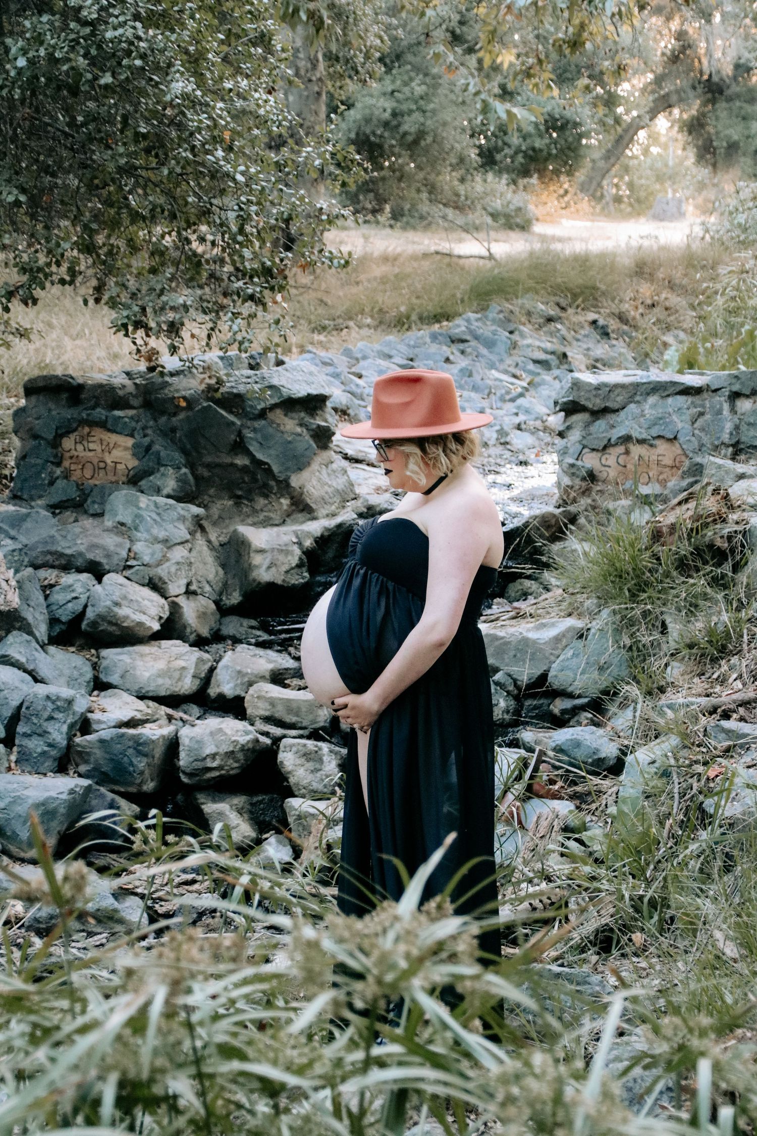 Person in dark dress and brown hat explores rocky terrain amidst wild grasses and stone walls.