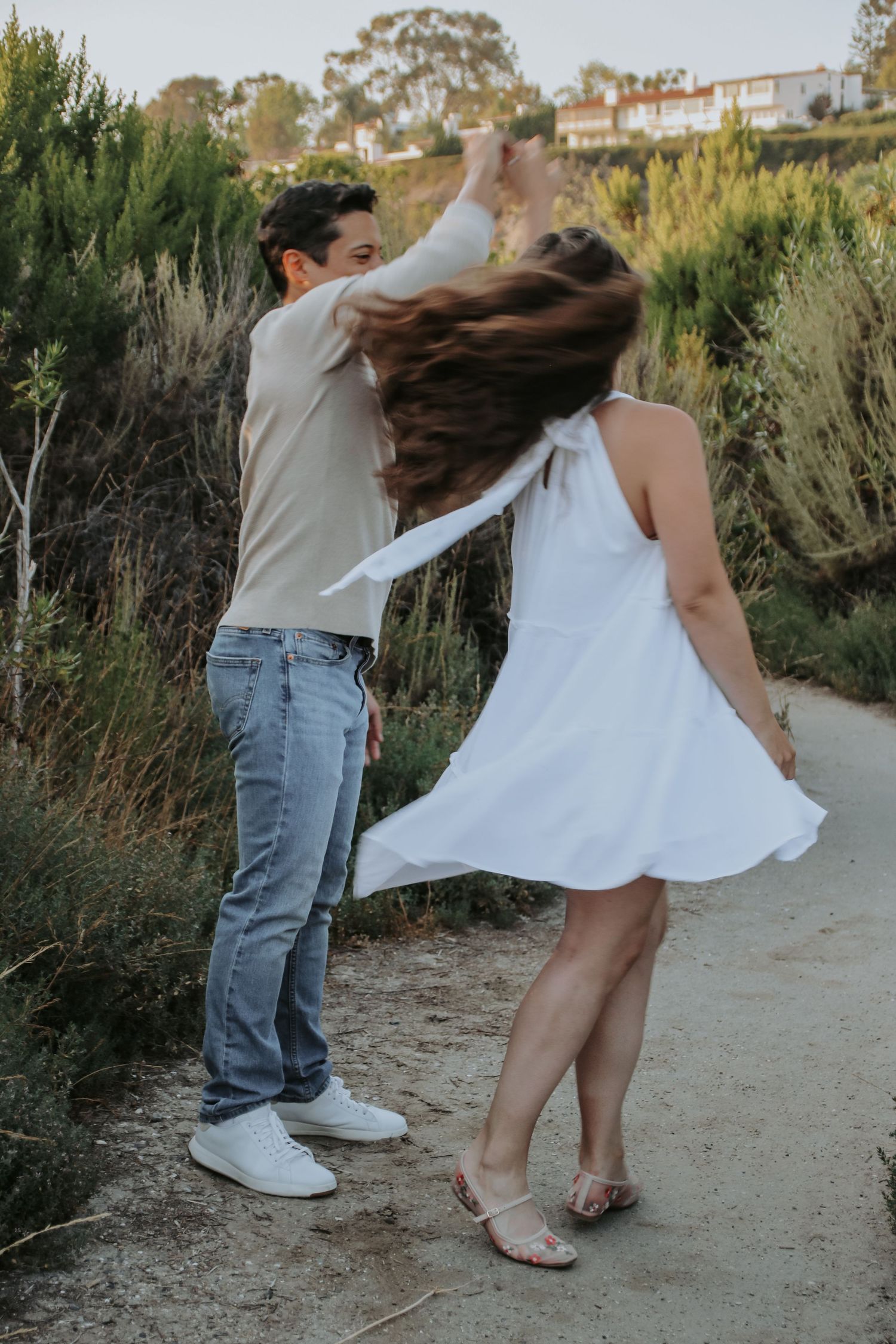 A couple dances playfully on a coastal path with wild sage brush and hillside homes in the background.