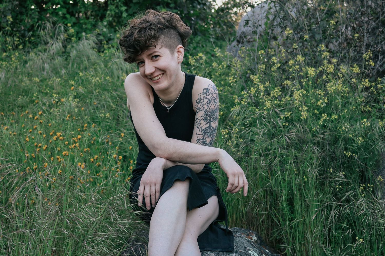 Portrait in wildflower meadow shows relaxed pose with black sleeveless top and arm tattoo.
