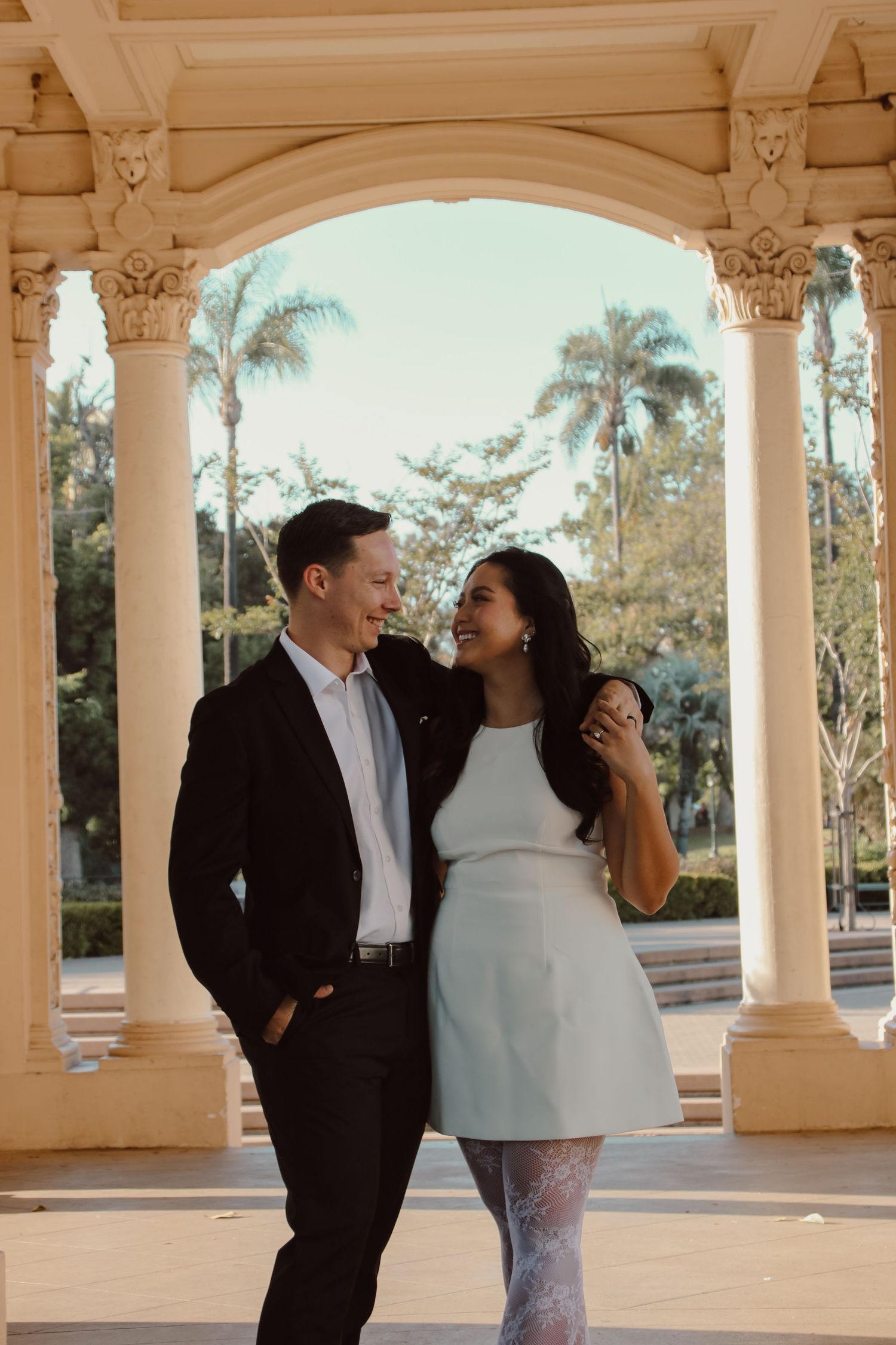 Two people share joyful moments in a colonnade with palm trees visible through arches.