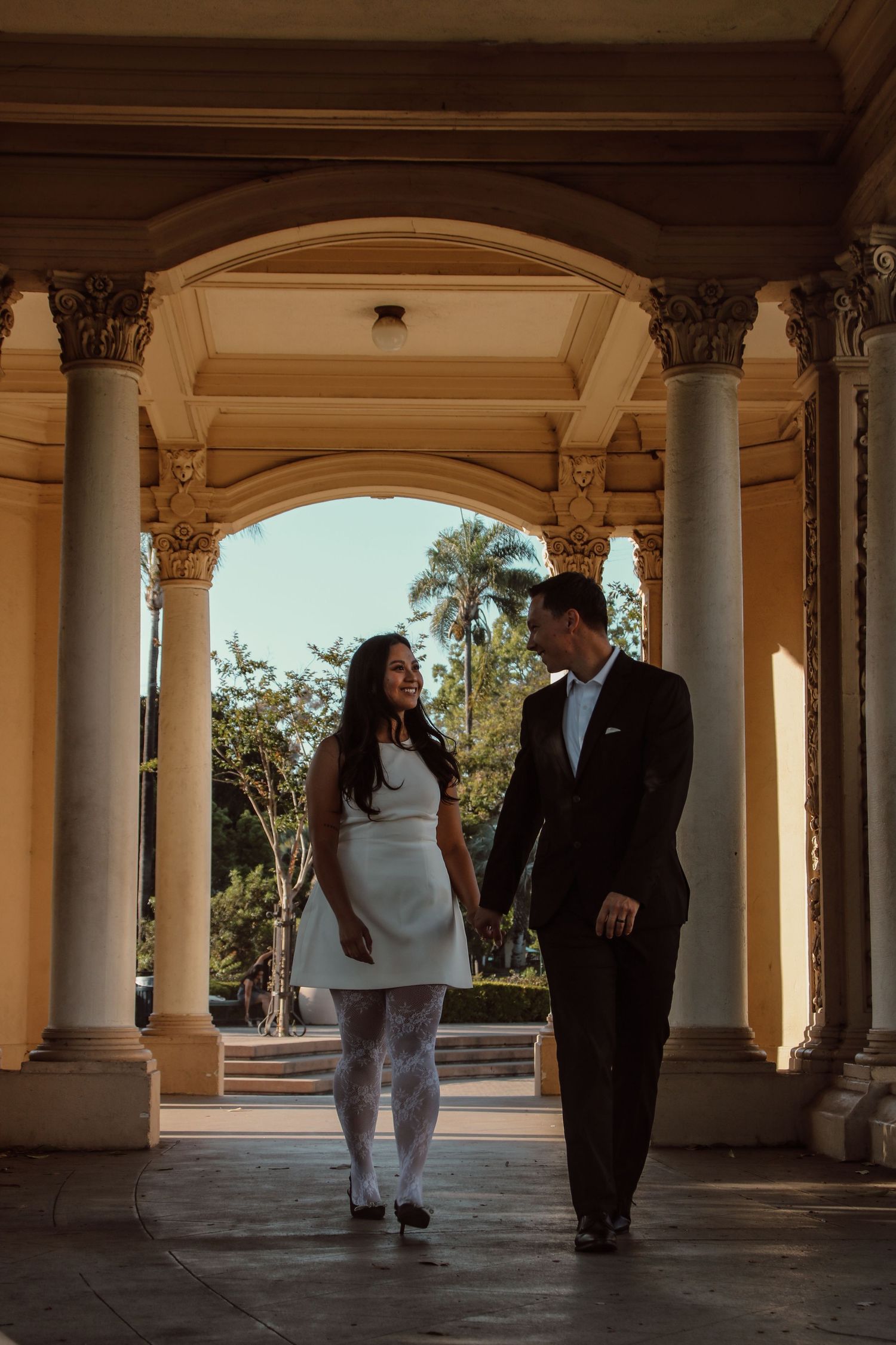 Romantic couple enjoying moment together in sunlit colonnade with ornate pillars.