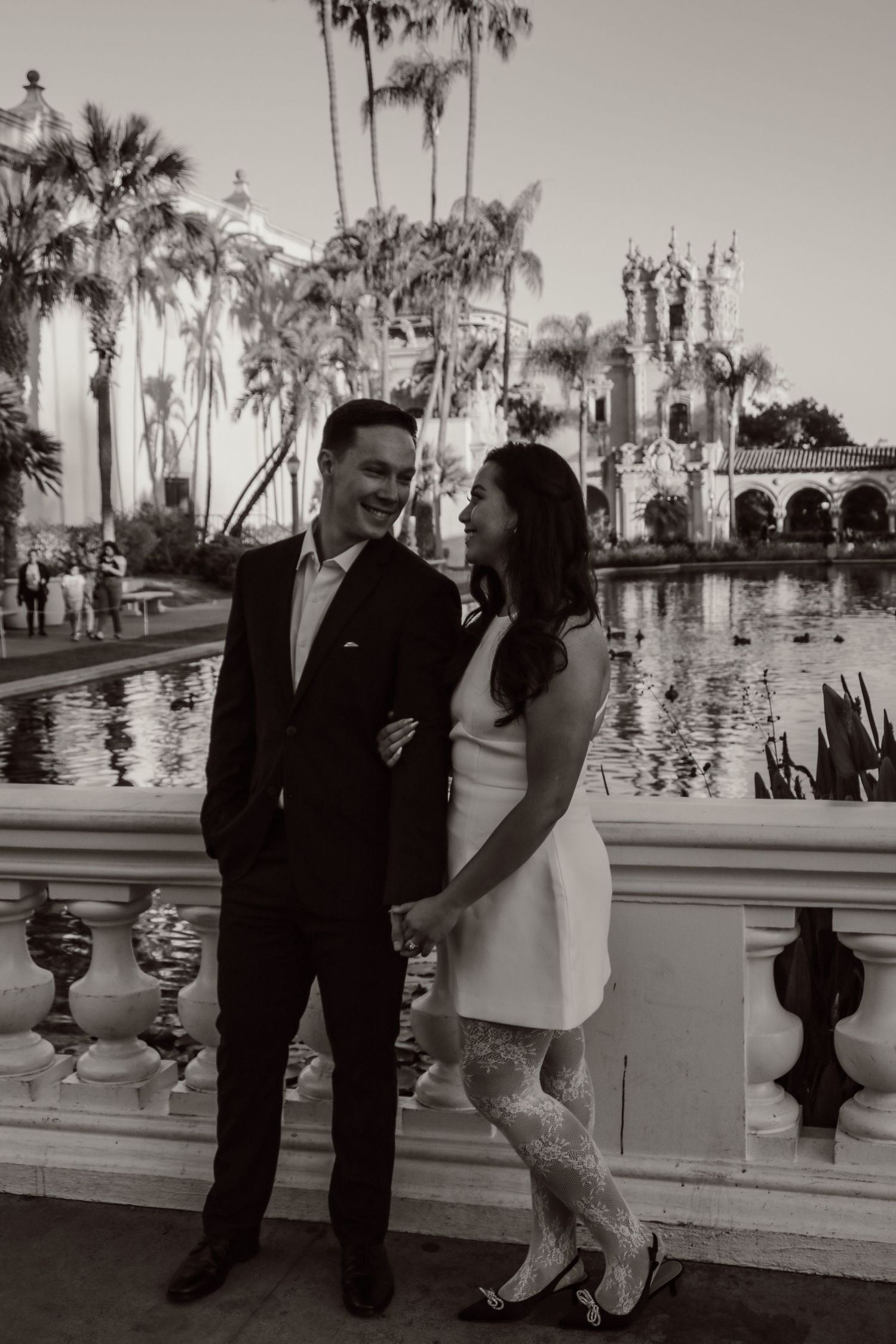 Black and white portrait of couple by reflecting pool with Spanish colonial architecture and palm trees.