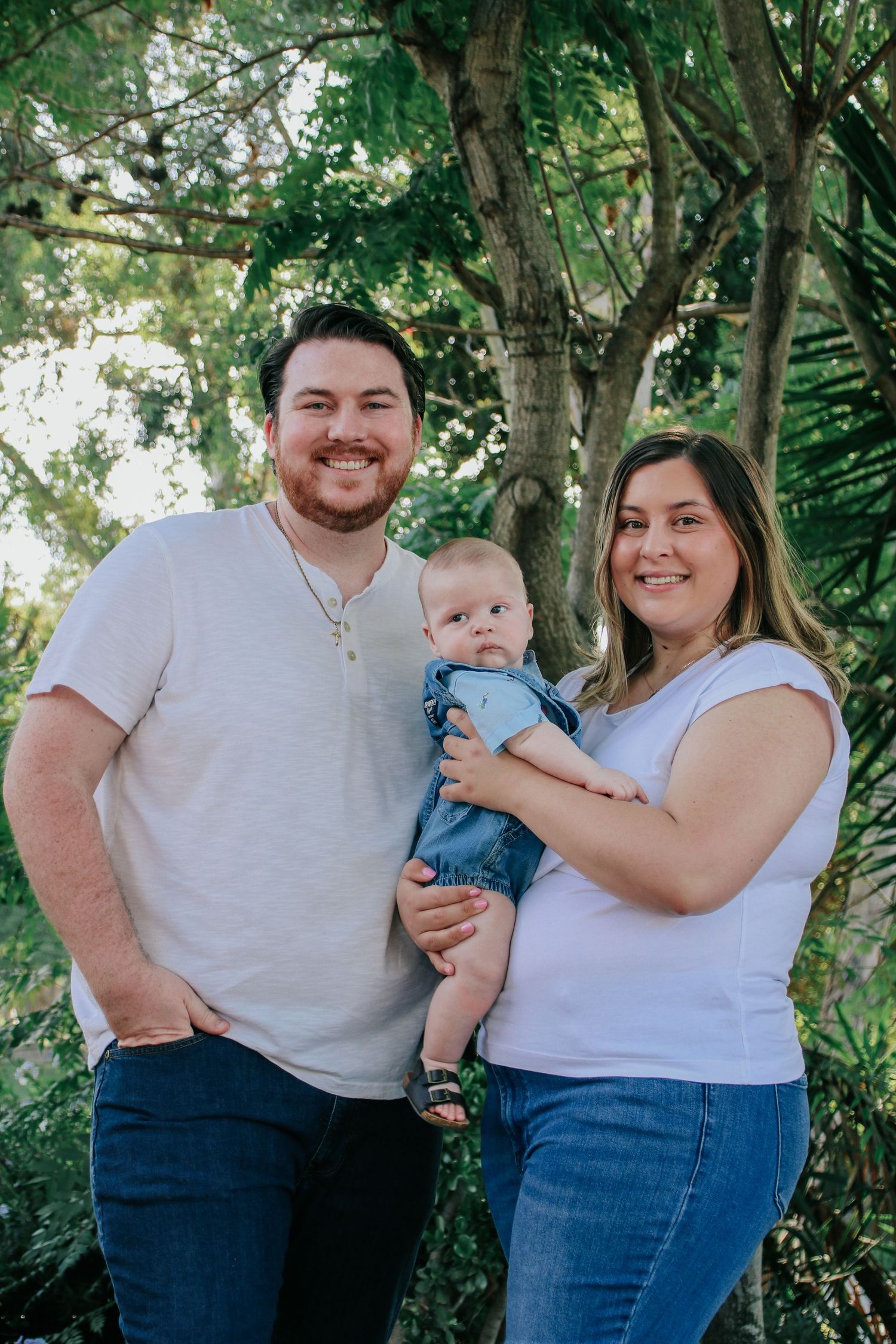 Family of three poses in casual attire against greenery in outdoor portrait session.