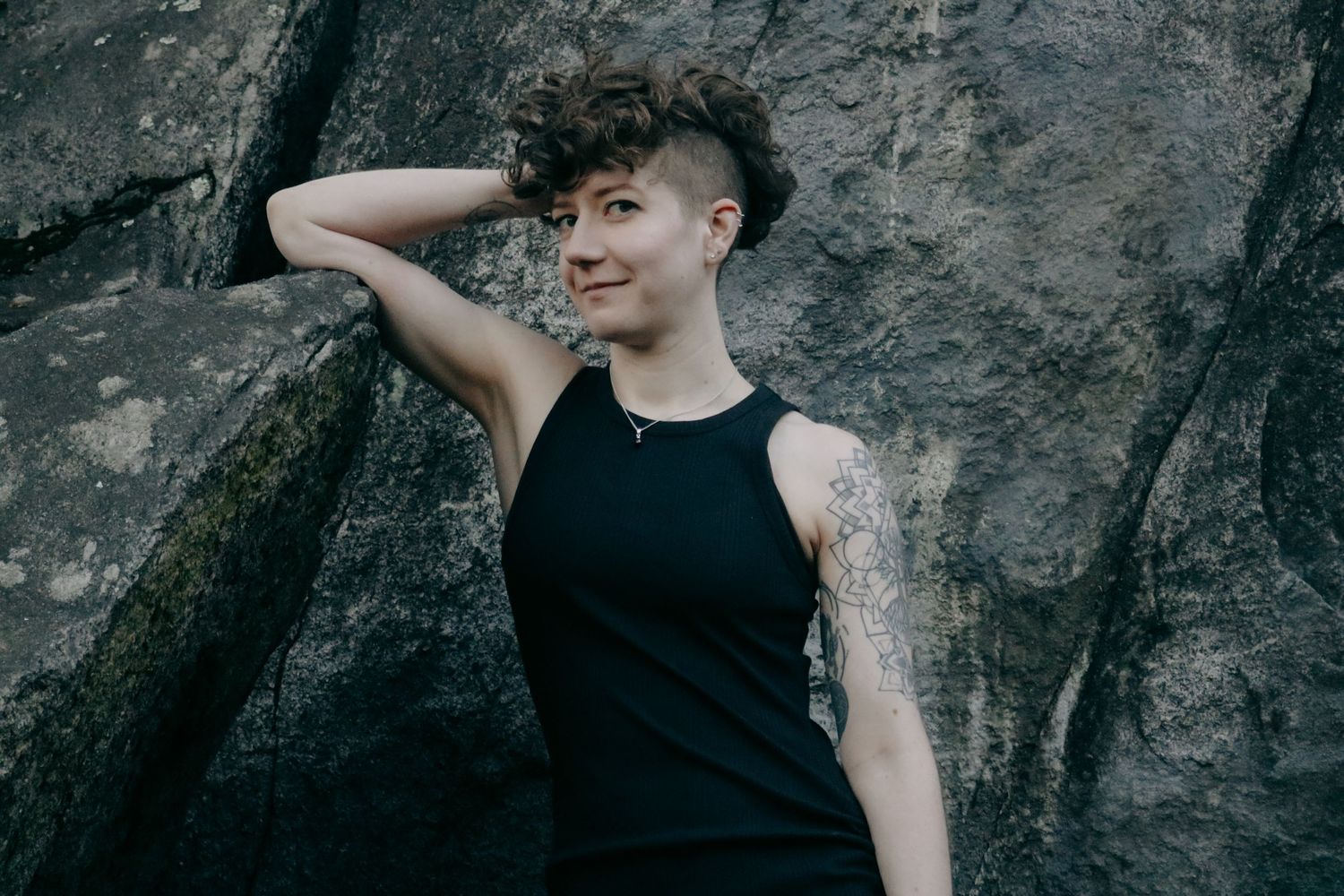Person with short dark hair and tattoo sleeve poses confidently against rocky wall in black tank top.