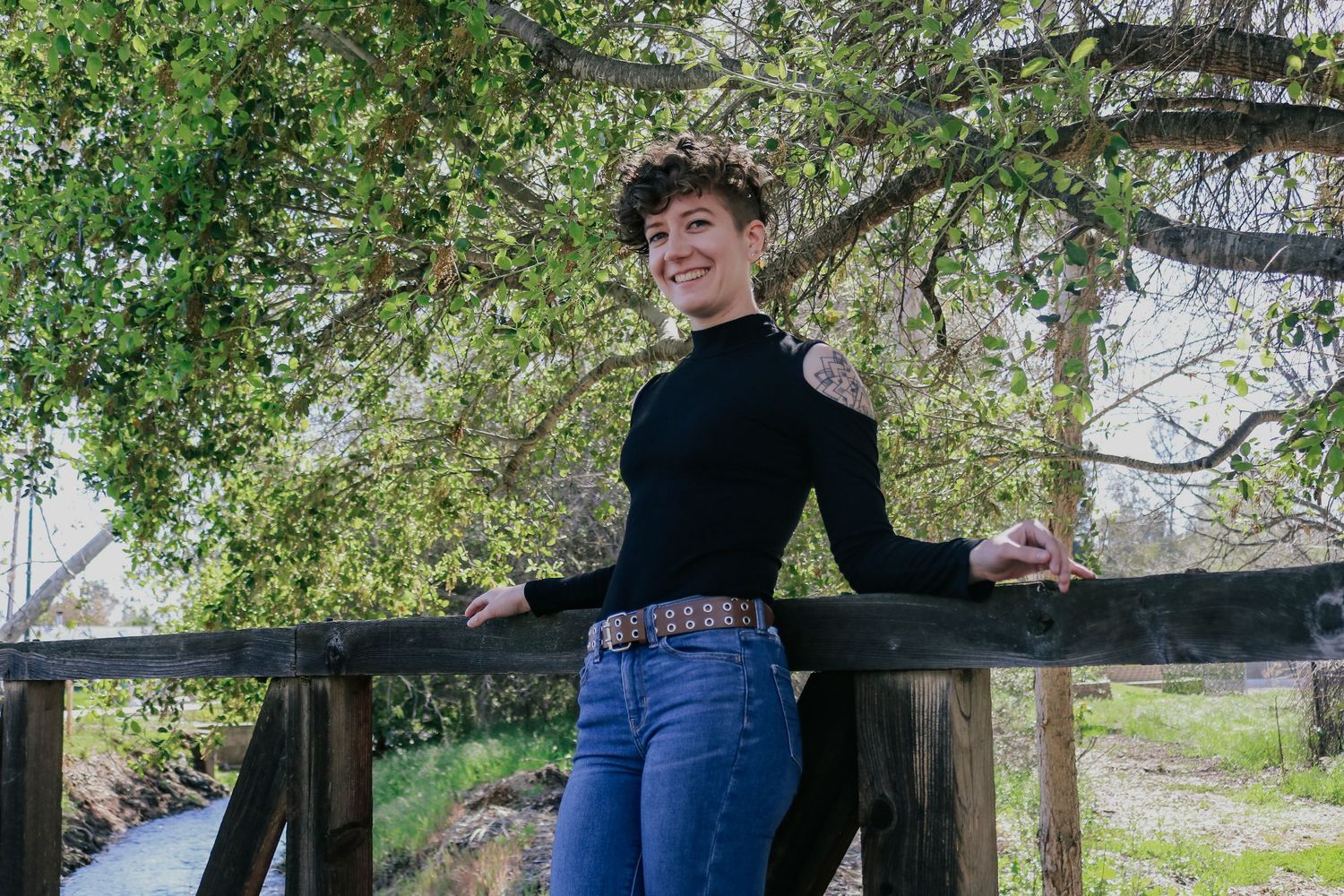 A person in a black turtleneck and jeans poses casually against a wooden railing in an outdoor nature setting.