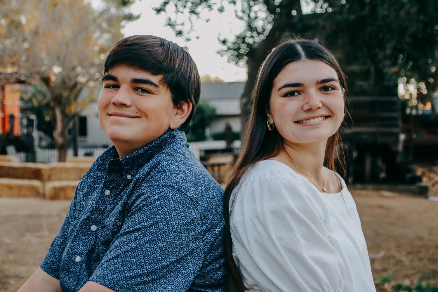 Casual outdoor portrait of two people sitting together with stone wall and trees in background.