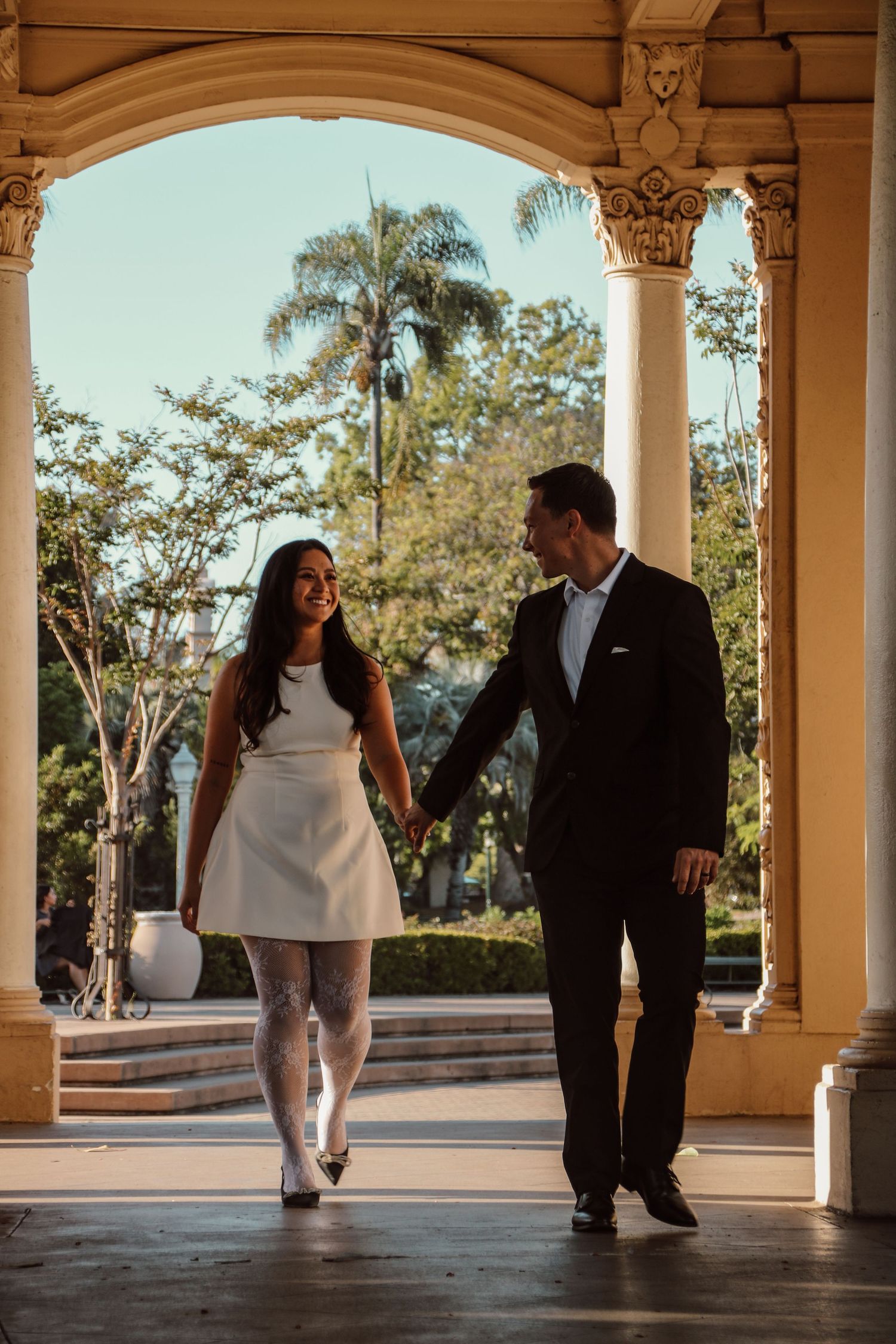 A couple walks hand in hand under ornate columns during a romantic outdoor photoshoot.