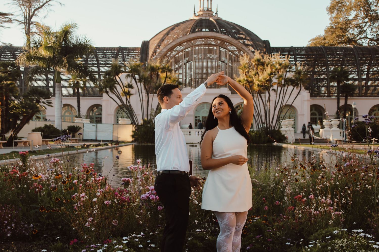 A joyful dance pose captured in front of an ornate botanical building with landscaped gardens.