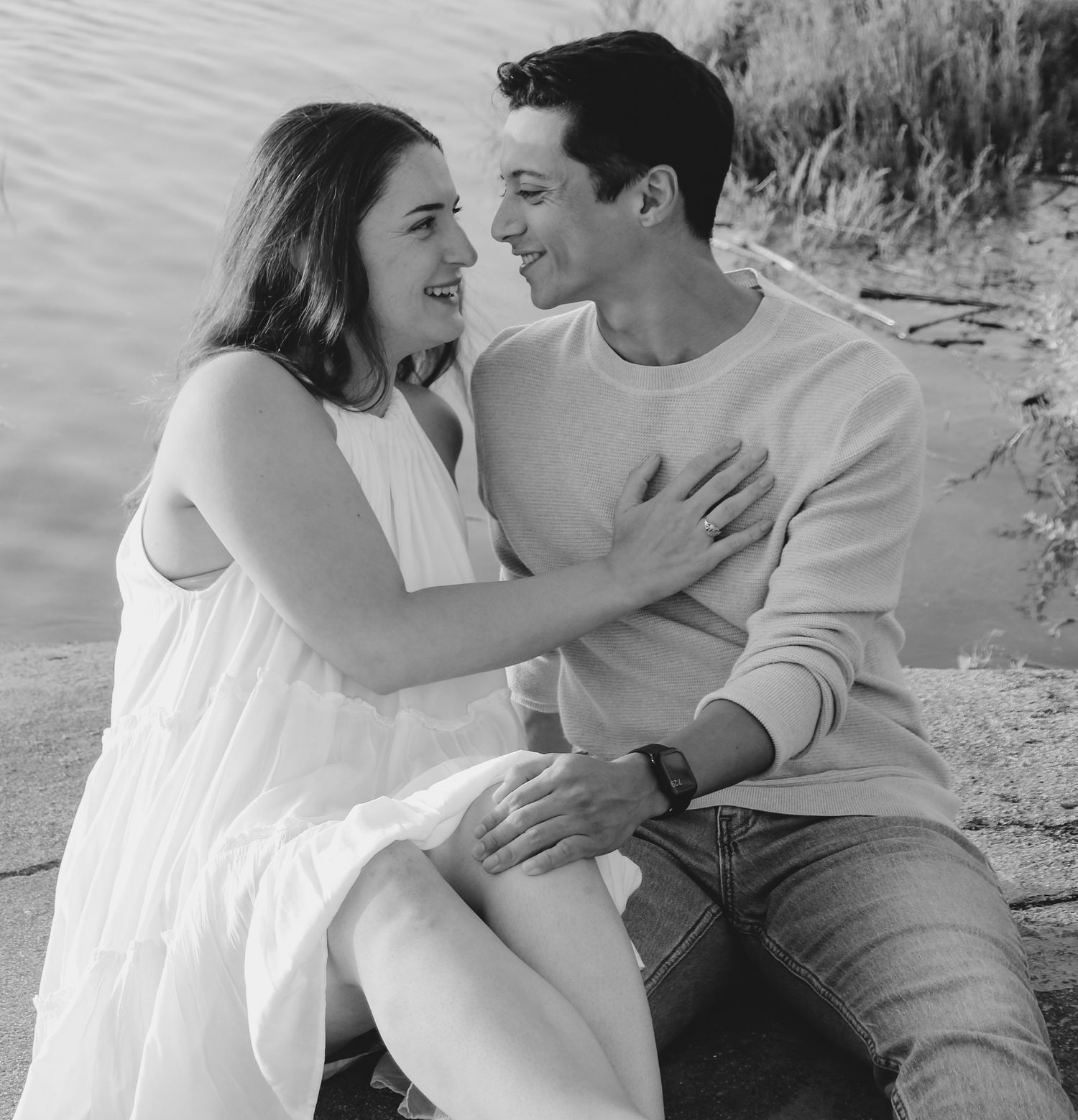 Romantic couple sharing an intimate moment together while seated on rocks by the water in black and white.