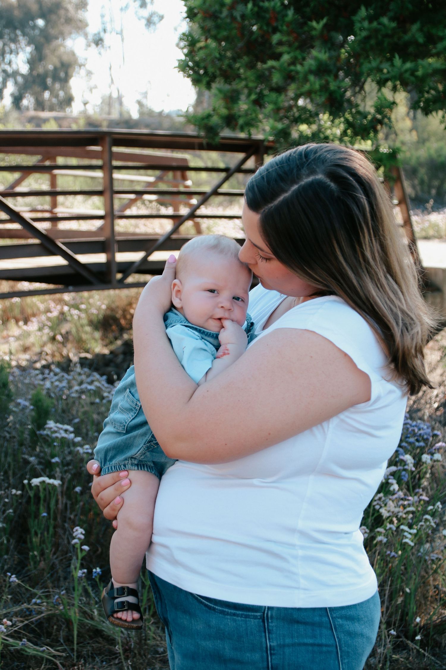 Mother in white shirt holding infant in blue outfit against nature backdrop with wooden fence.