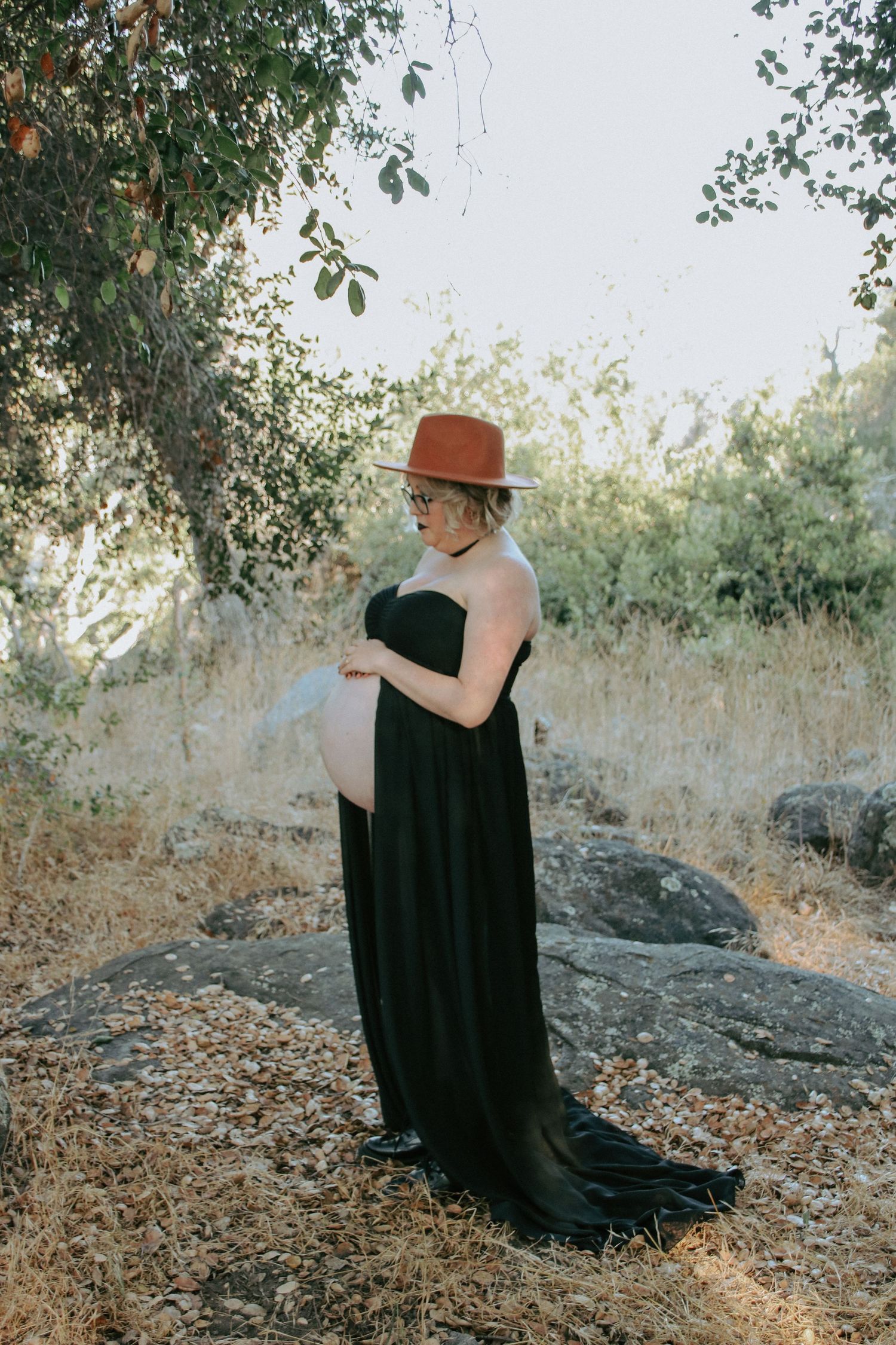 Individual in a long black dress sits contemplatively on rocks surrounded by trees and foliage.