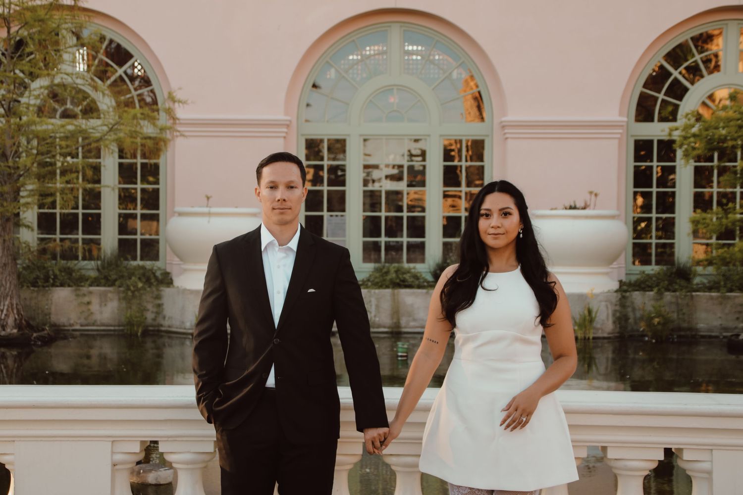 Stylish couple in formal wear pose together against ornate building with arched windows.