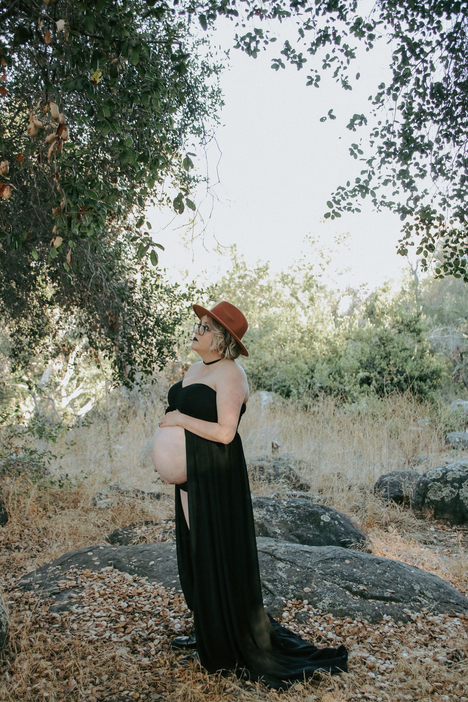 Maternity photoshoot in nature featuring a flowing black dress and brown hat against a backdrop of trees and rocks.