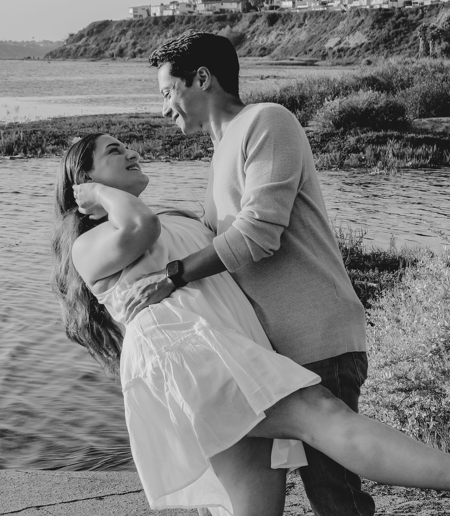 Playful dip pose on waterfront with cliffs in background during romantic photoshoot.