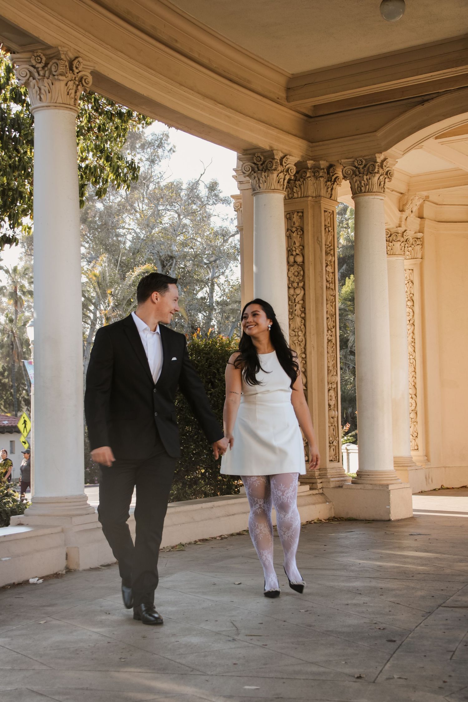 Two people share laughter while walking through an elegant colonnade with ornate architectural details.