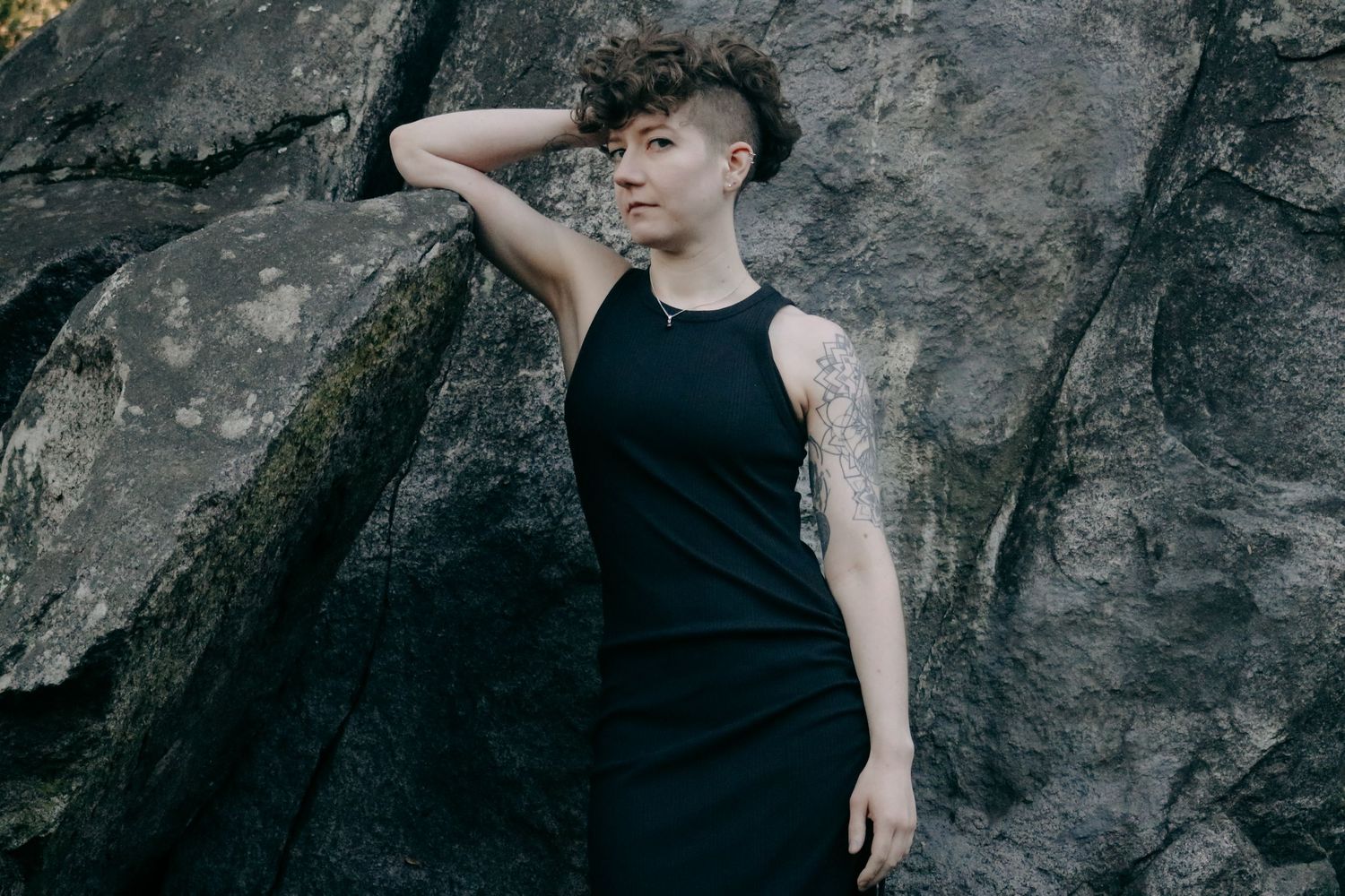 Moody outdoor portrait against stone wall showing person in black sleeveless dress with arm raised.