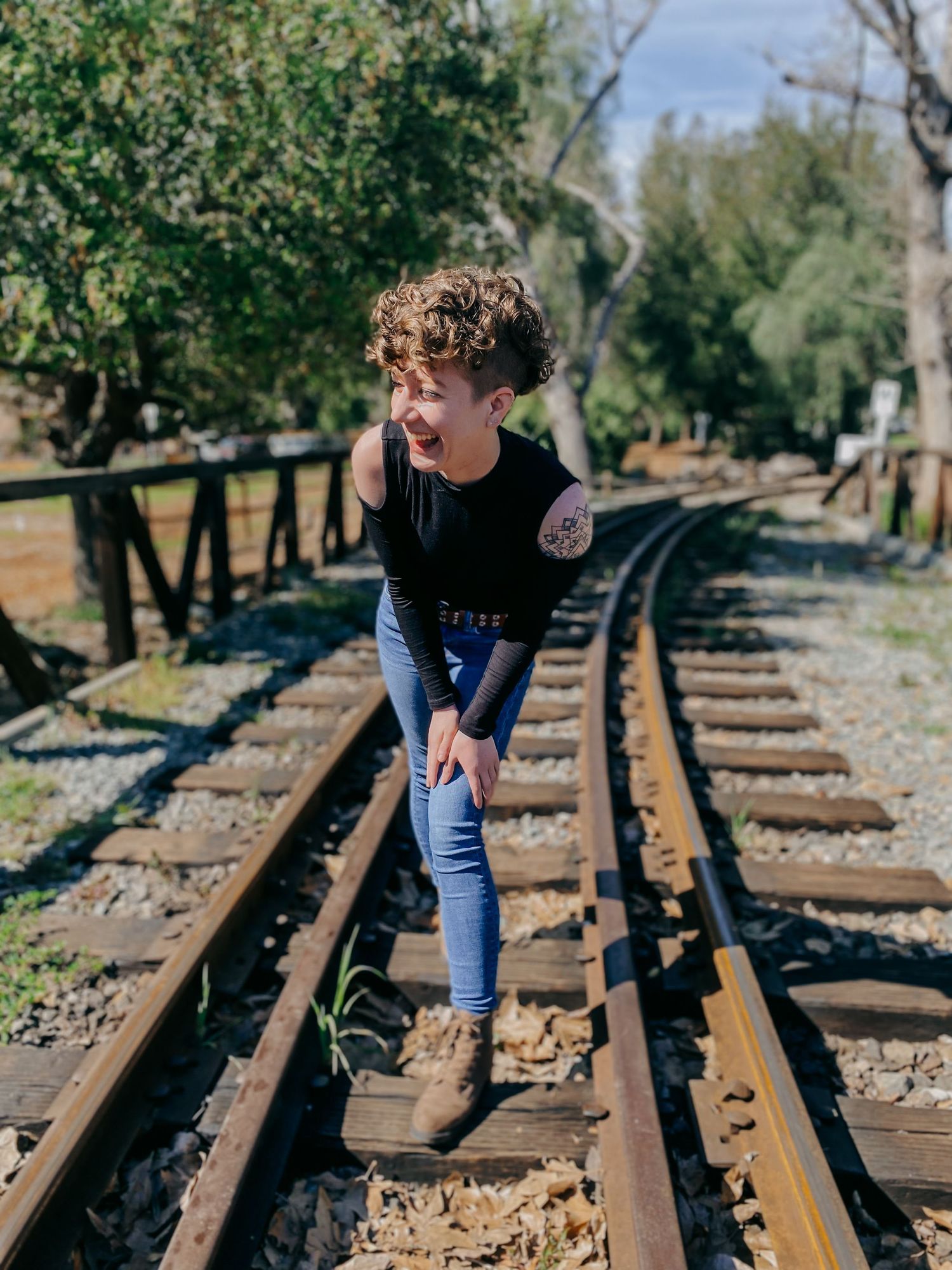 A person balances on railroad tracks while wearing a black top and jeans in a wooded setting.