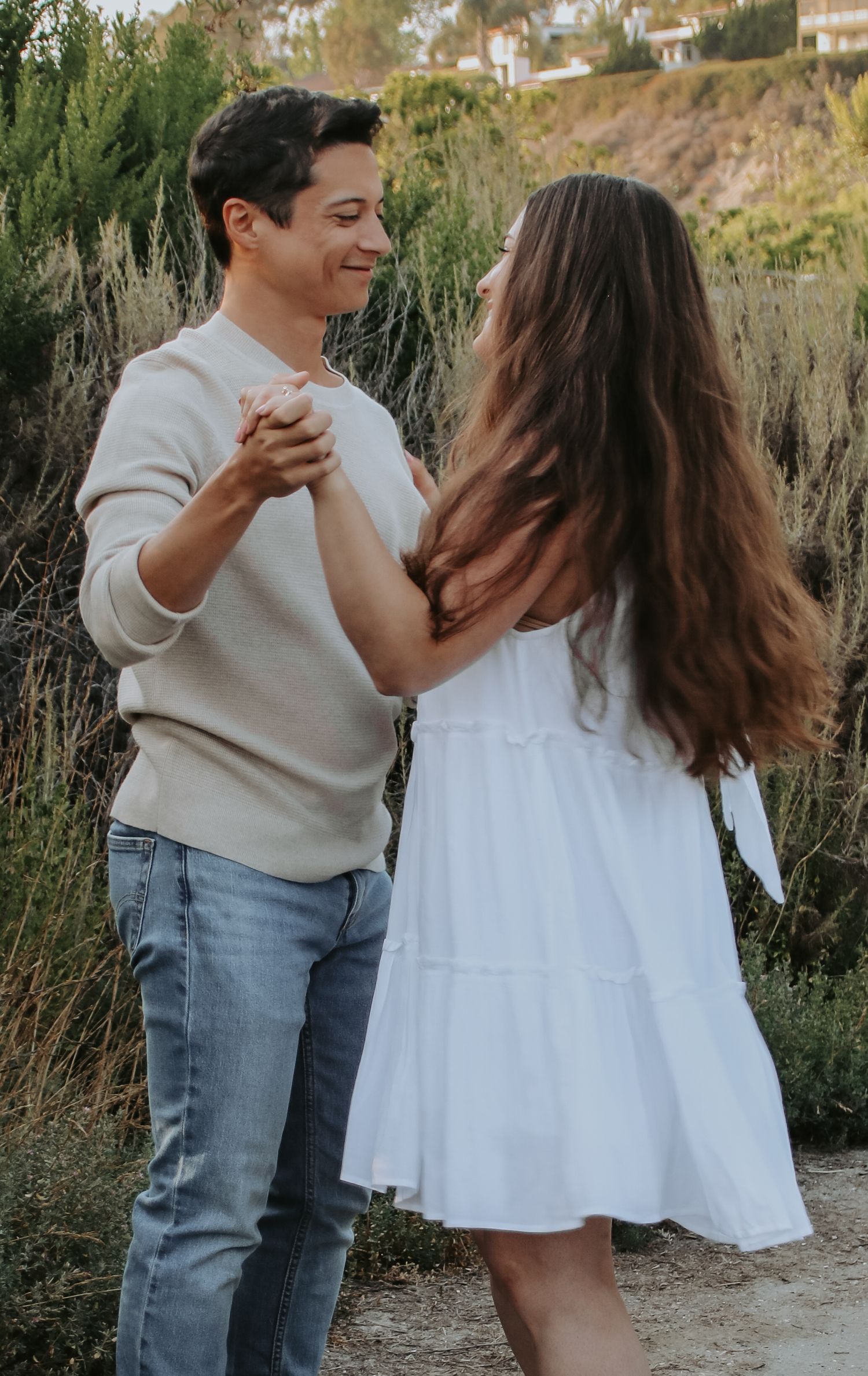 A couple holds hands while dancing on a dirt path surrounded by coastal vegetation.