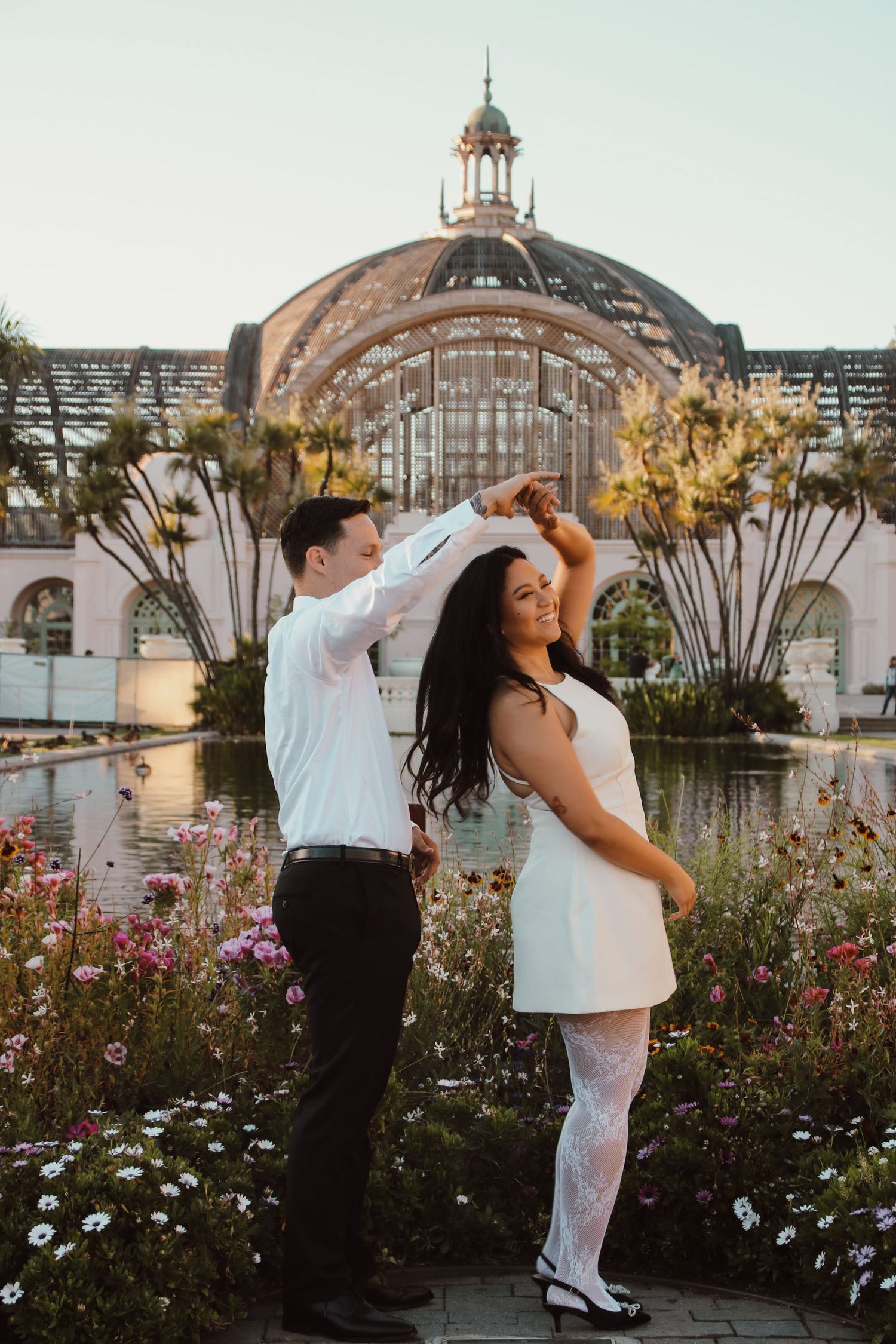 A playful dancing pose in front of a grand glass-domed conservatory with reflecting pools and gardens.