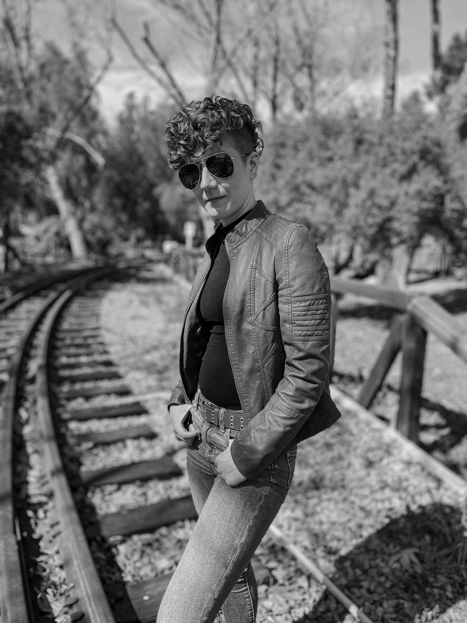Black and white photo of person in leather jacket posing beside vintage railway tracks.
