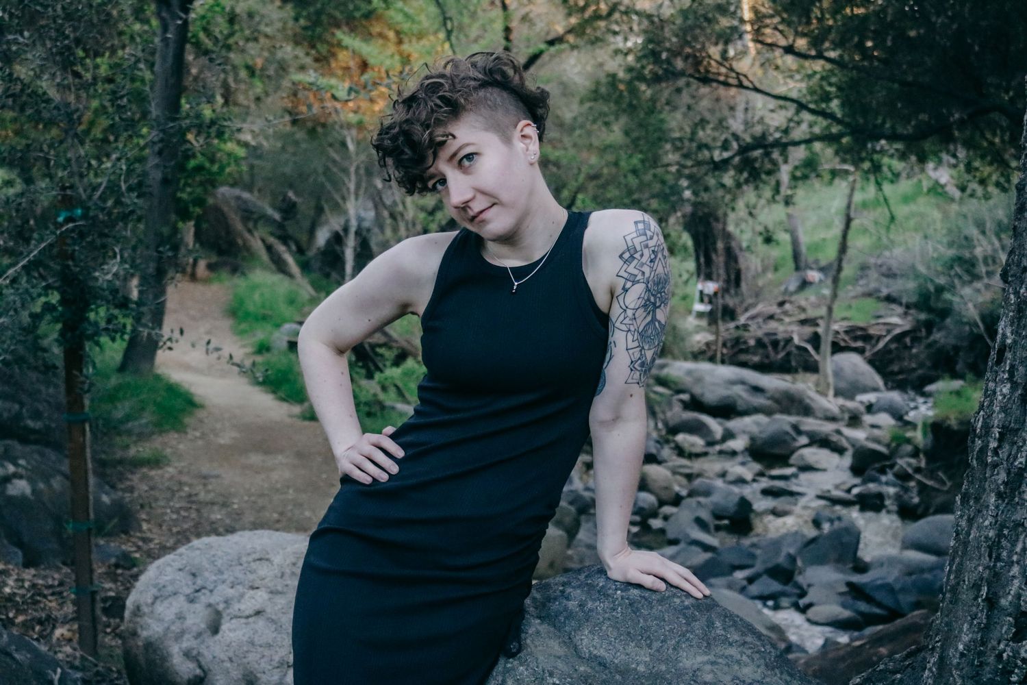 Person in black dress poses casually on rock formation near woodland stream.