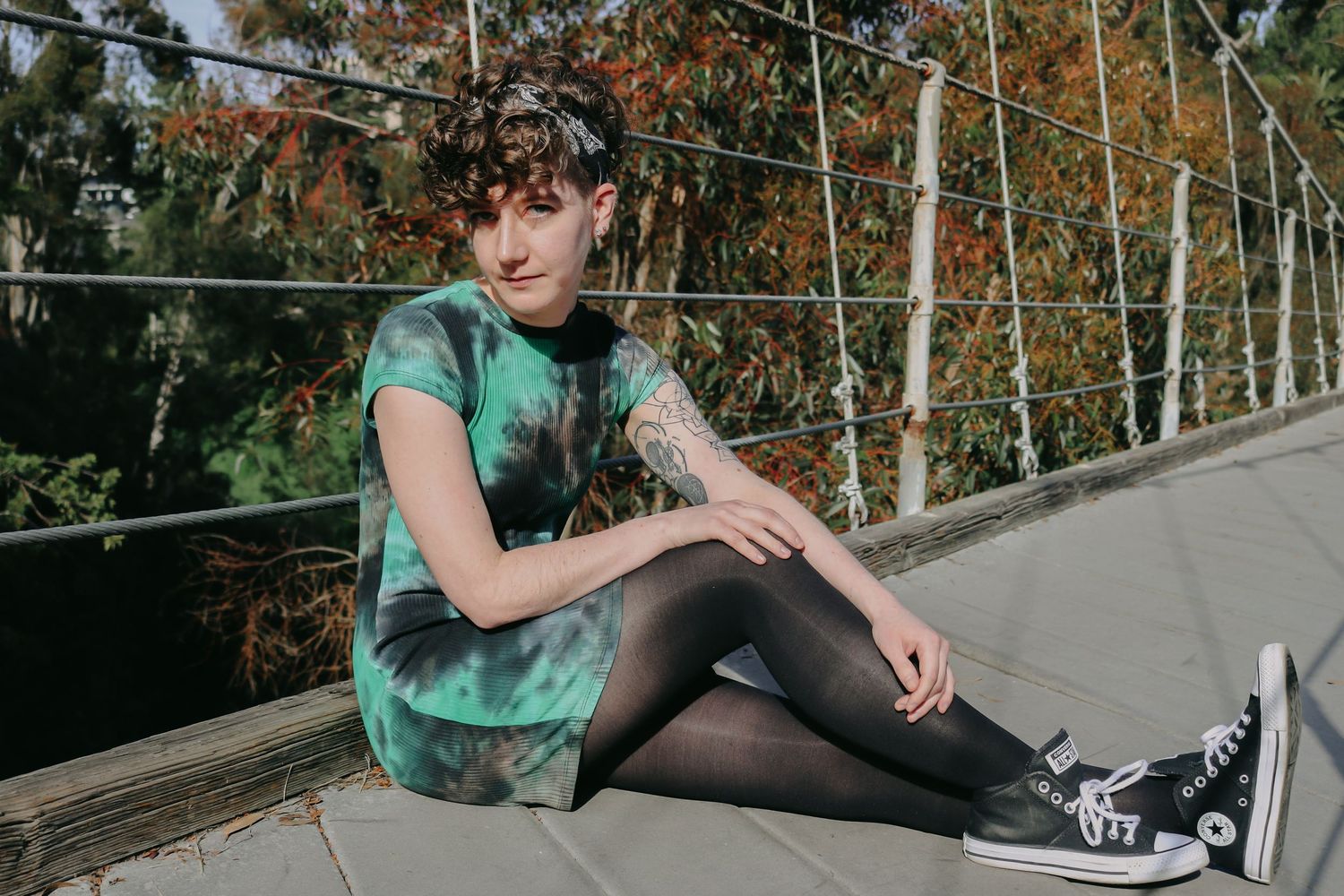 A person in a green tie-dye dress and tights sits on a bridge deck surrounded by fall foliage.