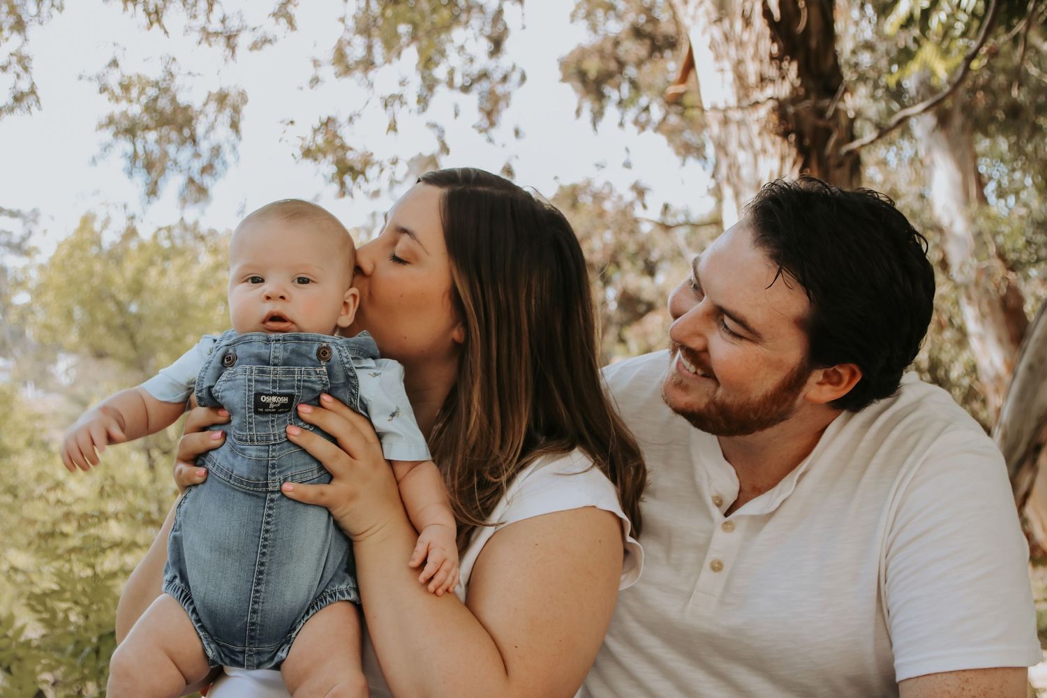 A warm family portrait showing loving moments between parents and their infant baby outdoors among trees.