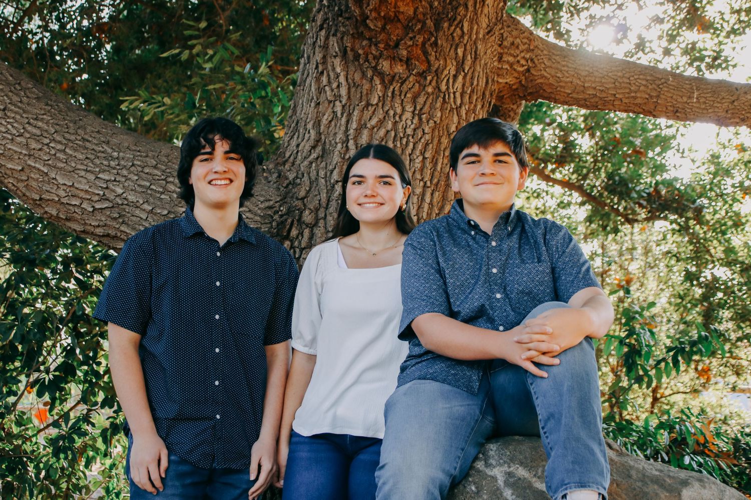 Three siblings pose casually together against tree trunk in outdoor setting.