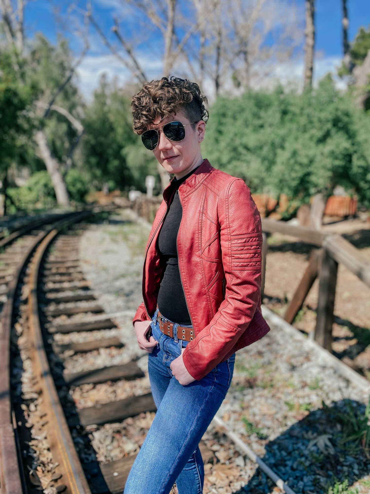 Person in red leather jacket and jeans poses along rustic railroad tracks surrounded by trees.
