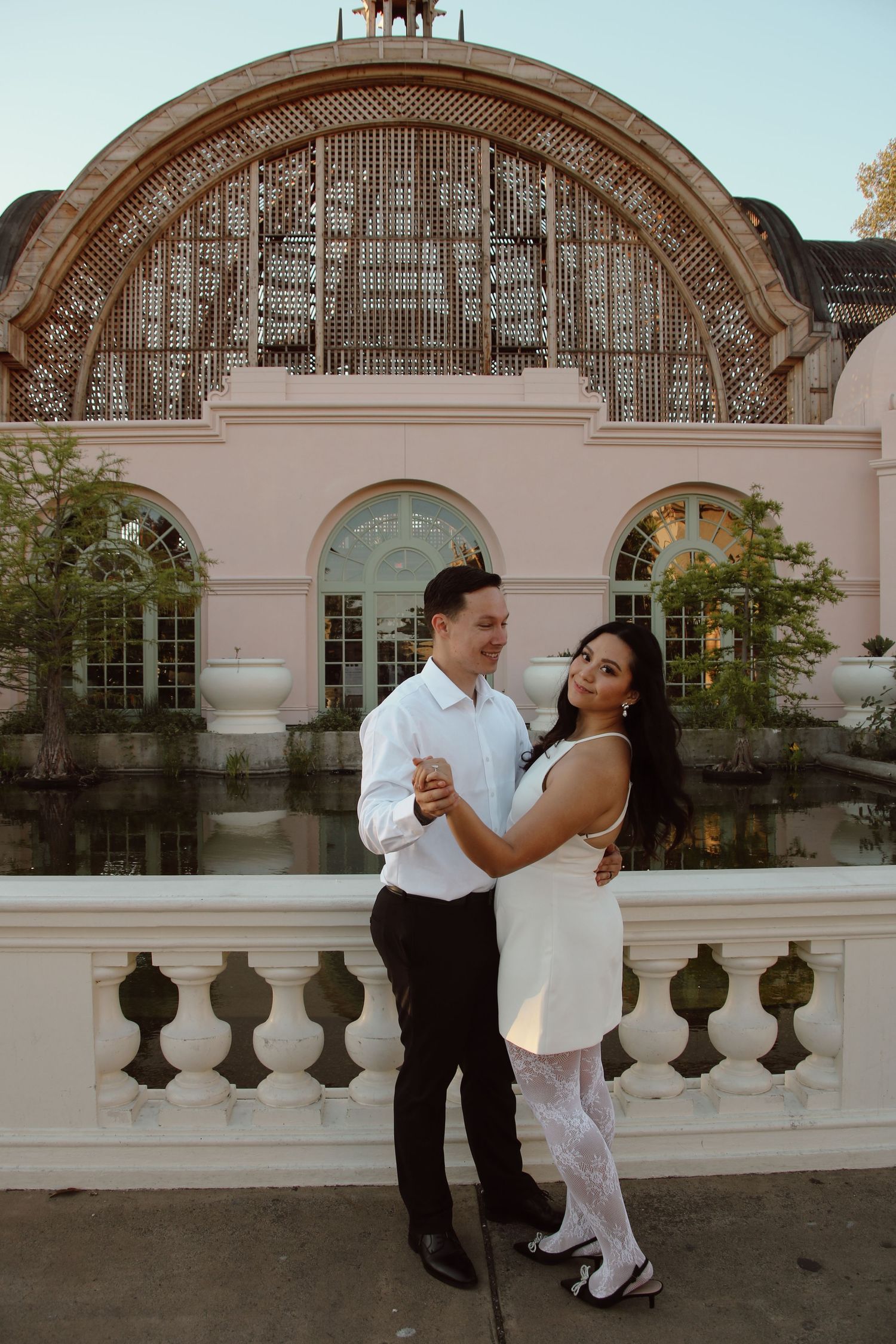 Dancing couple in coordinated black and white outfits posed by an elegant architectural balcony with water feature.