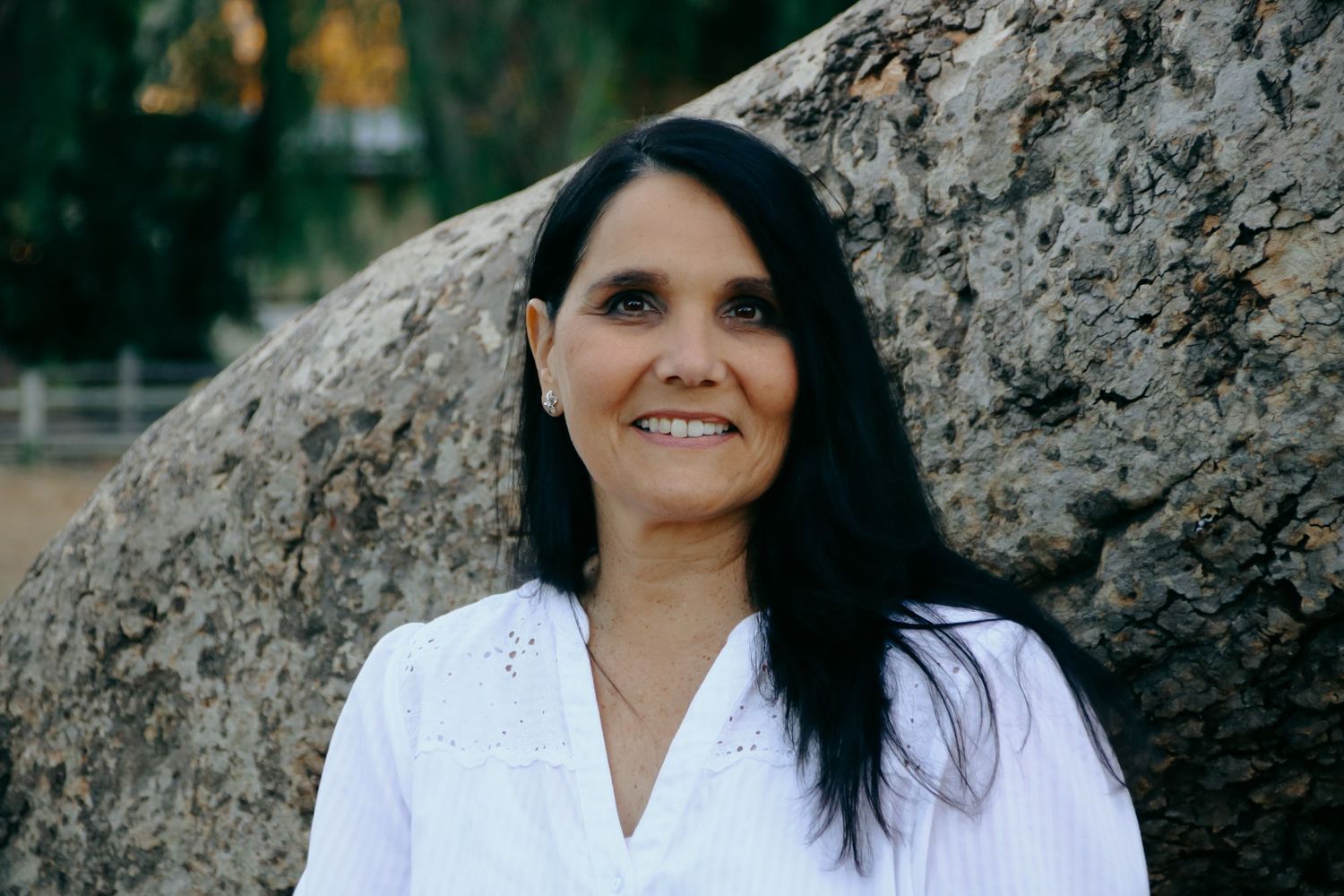 A person in a white top smiles while posing next to a large tree trunk outdoors.
