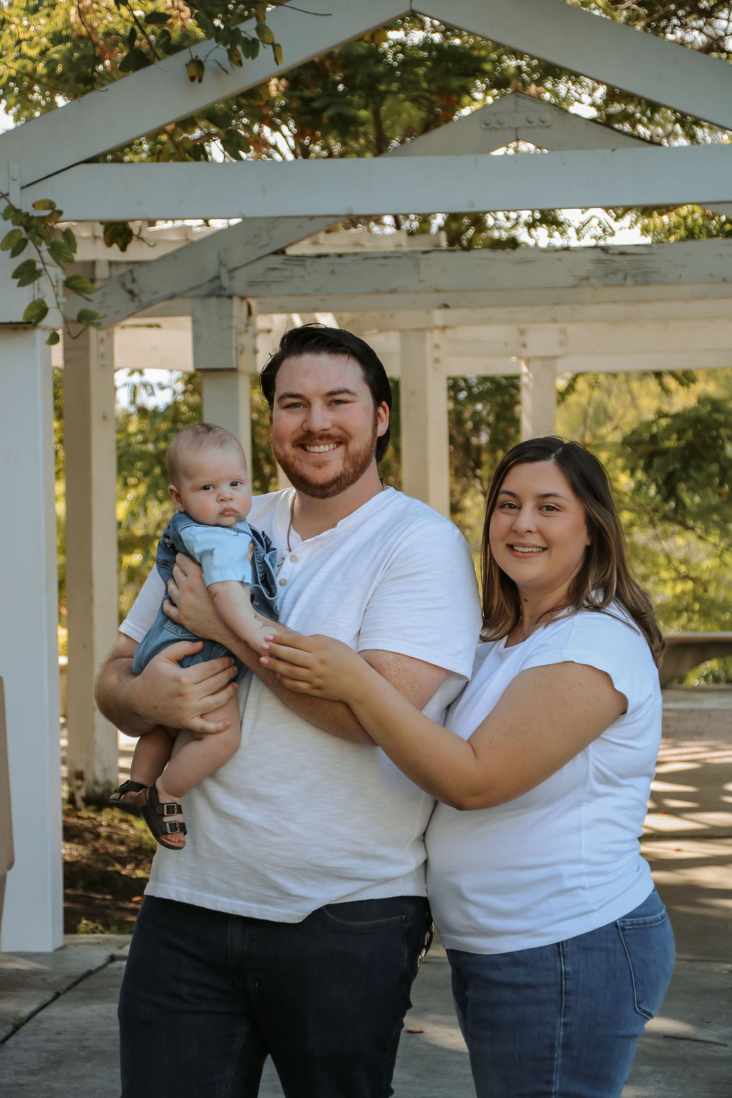 Smiling family poses with baby under white pergola during outdoor photo session.