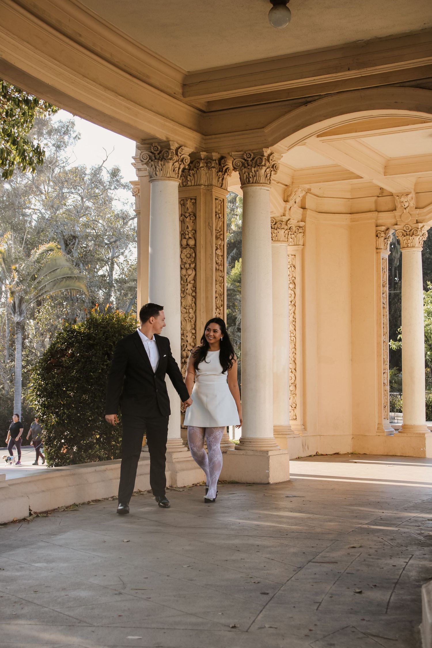 Two people walk and interact playfully in a classic columned corridor with natural light.