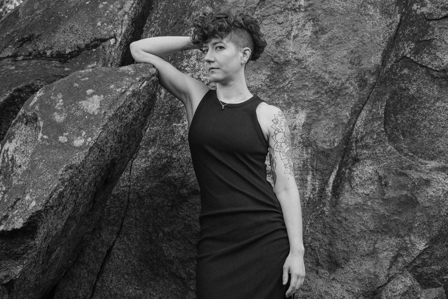 Figure in sleeveless black dress poses dramatically against rocky backdrop.