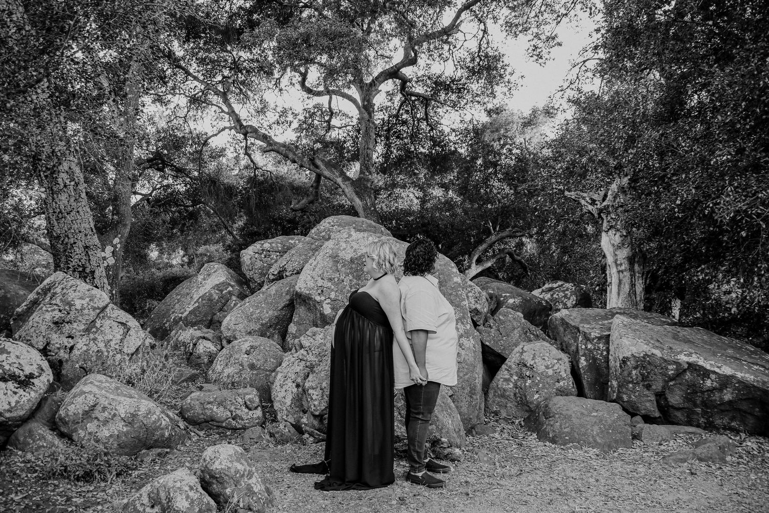 A couple stands together against large boulders and trees in a scenic black and white outdoor photo.