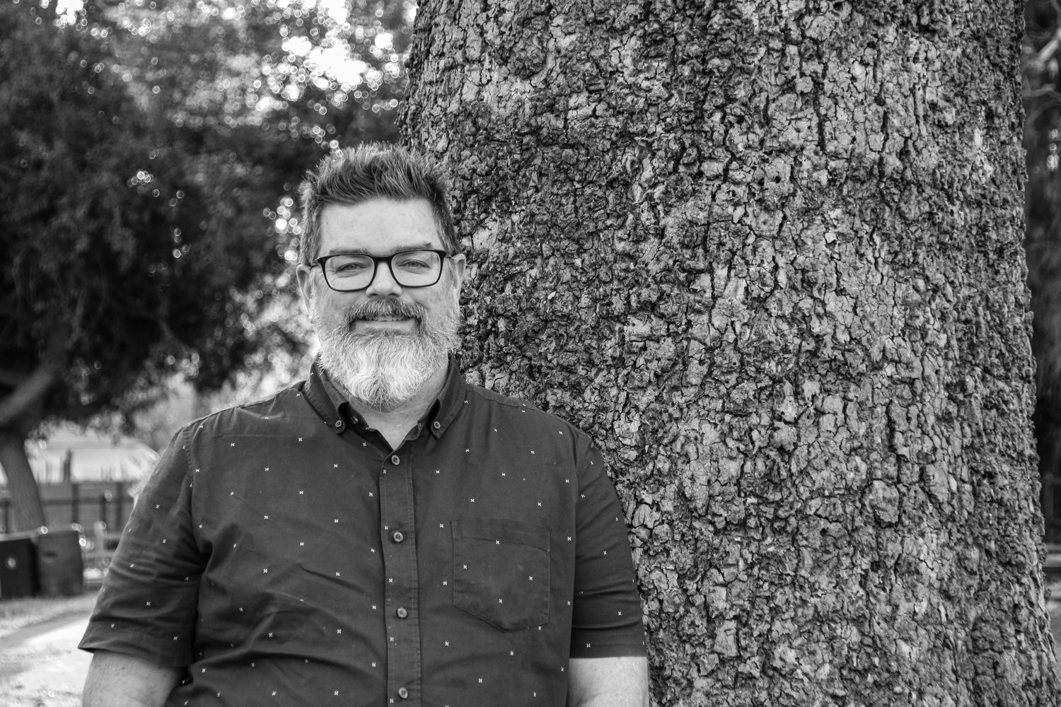 A bearded person in glasses and dark clothing poses next to a large tree trunk.