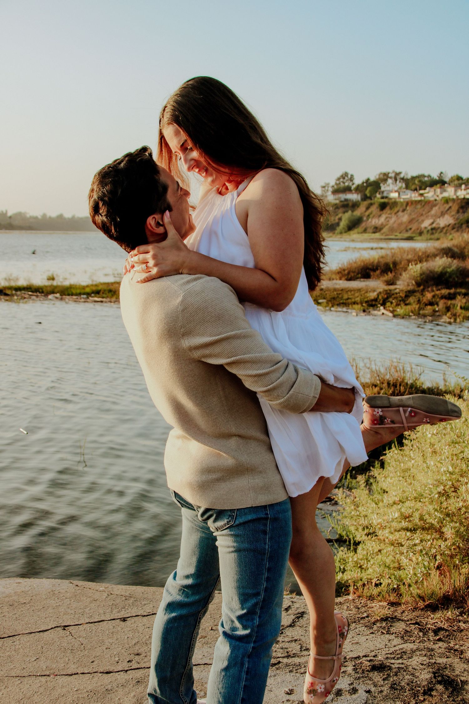 A couple embraces against a serene waterfront backdrop during sunset.