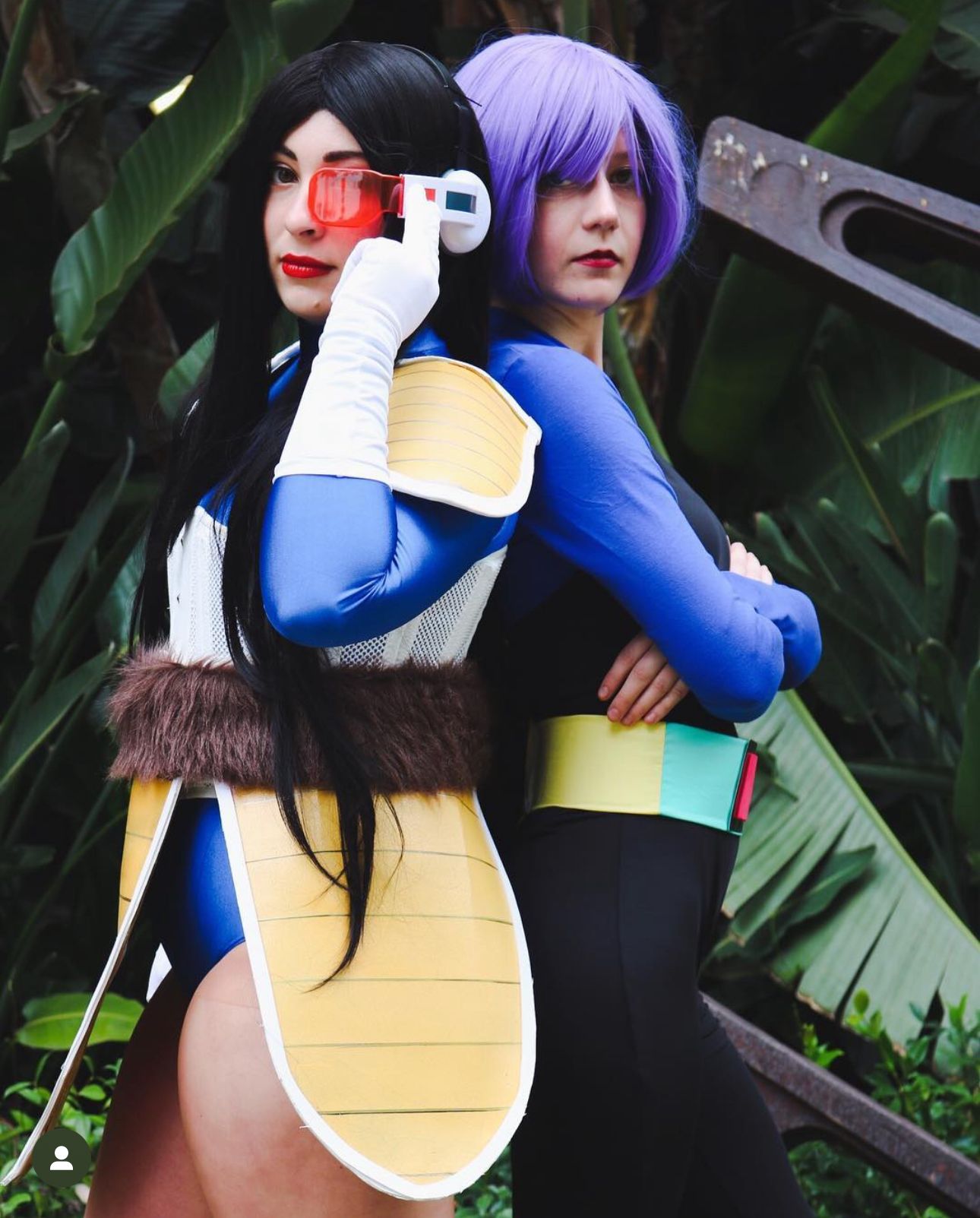 Two cosplayers in blue and yellow anime-inspired combat outfits pose together against foliage.