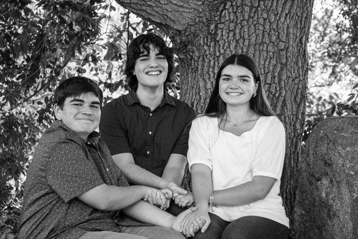 Black and white portrait of three siblings sitting together by large tree.