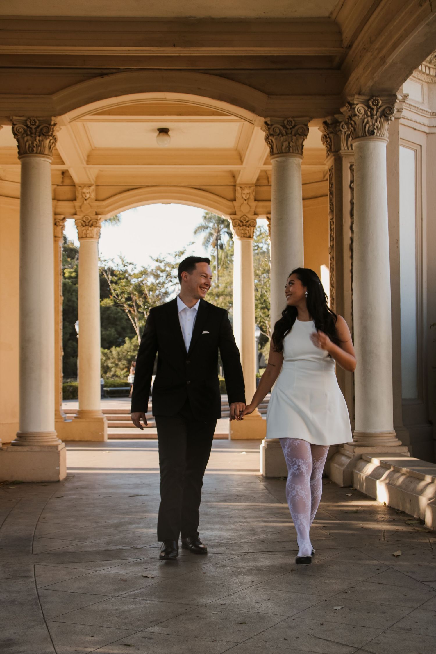 Elegant couple holding hands while walking through classical architecture with columns.