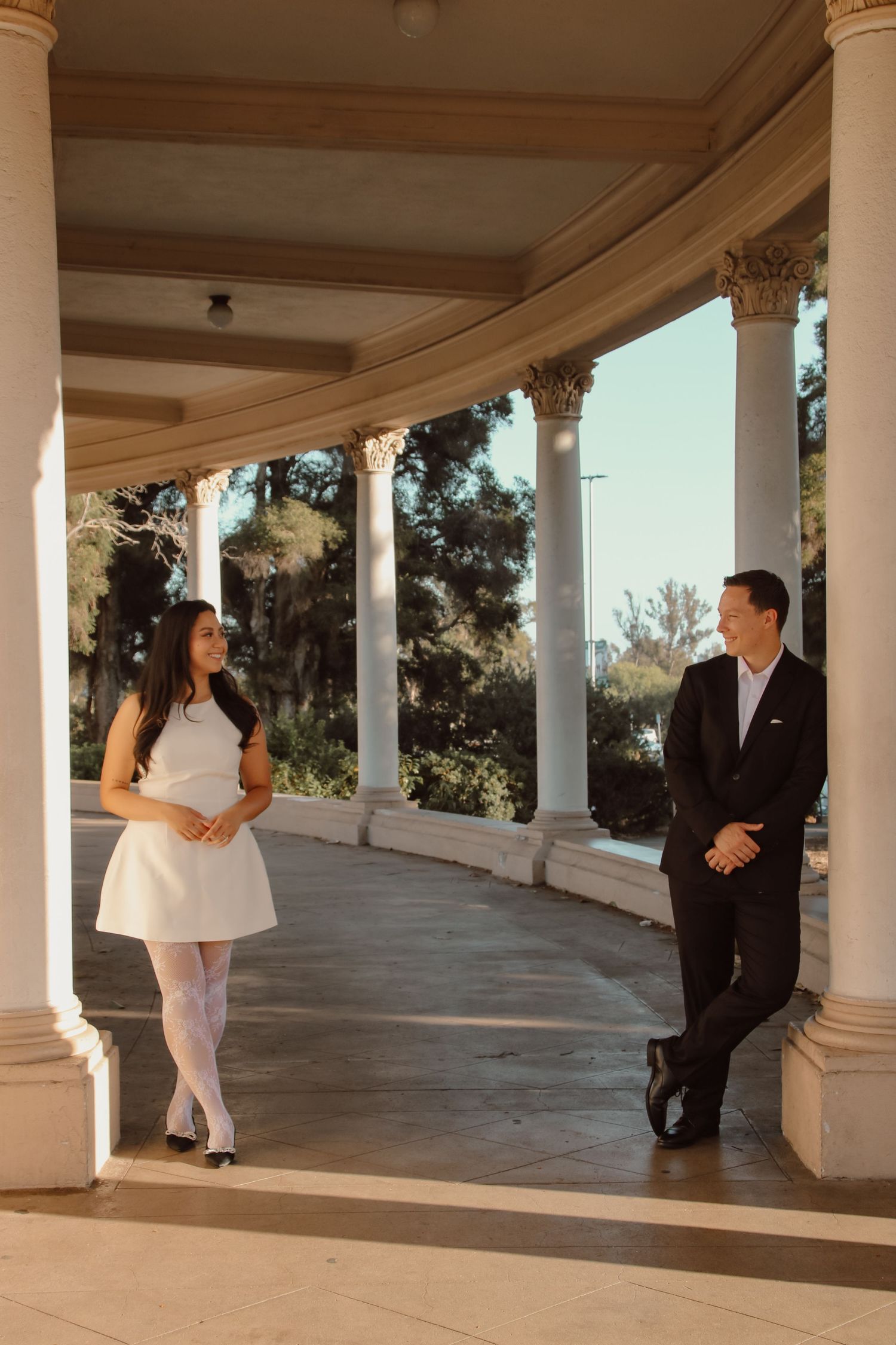 A couple stands at opposite ends of a curved colonnade with classical architecture.