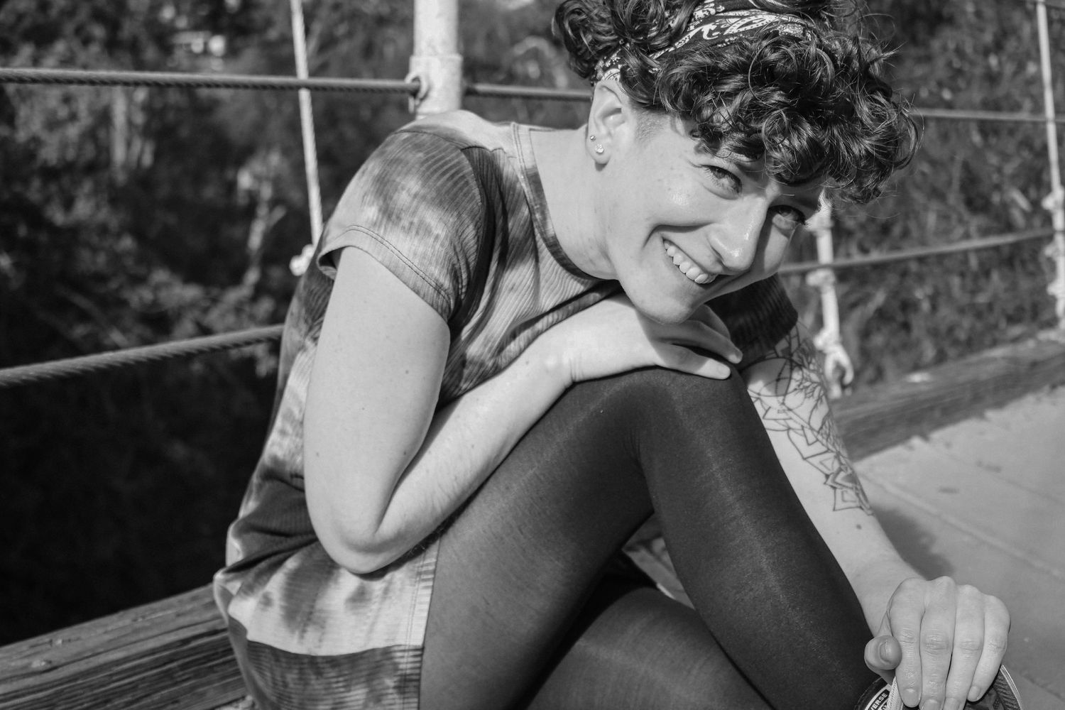 Person in black dress sits on rock in nature setting with curly hair and tattoo sleeve.