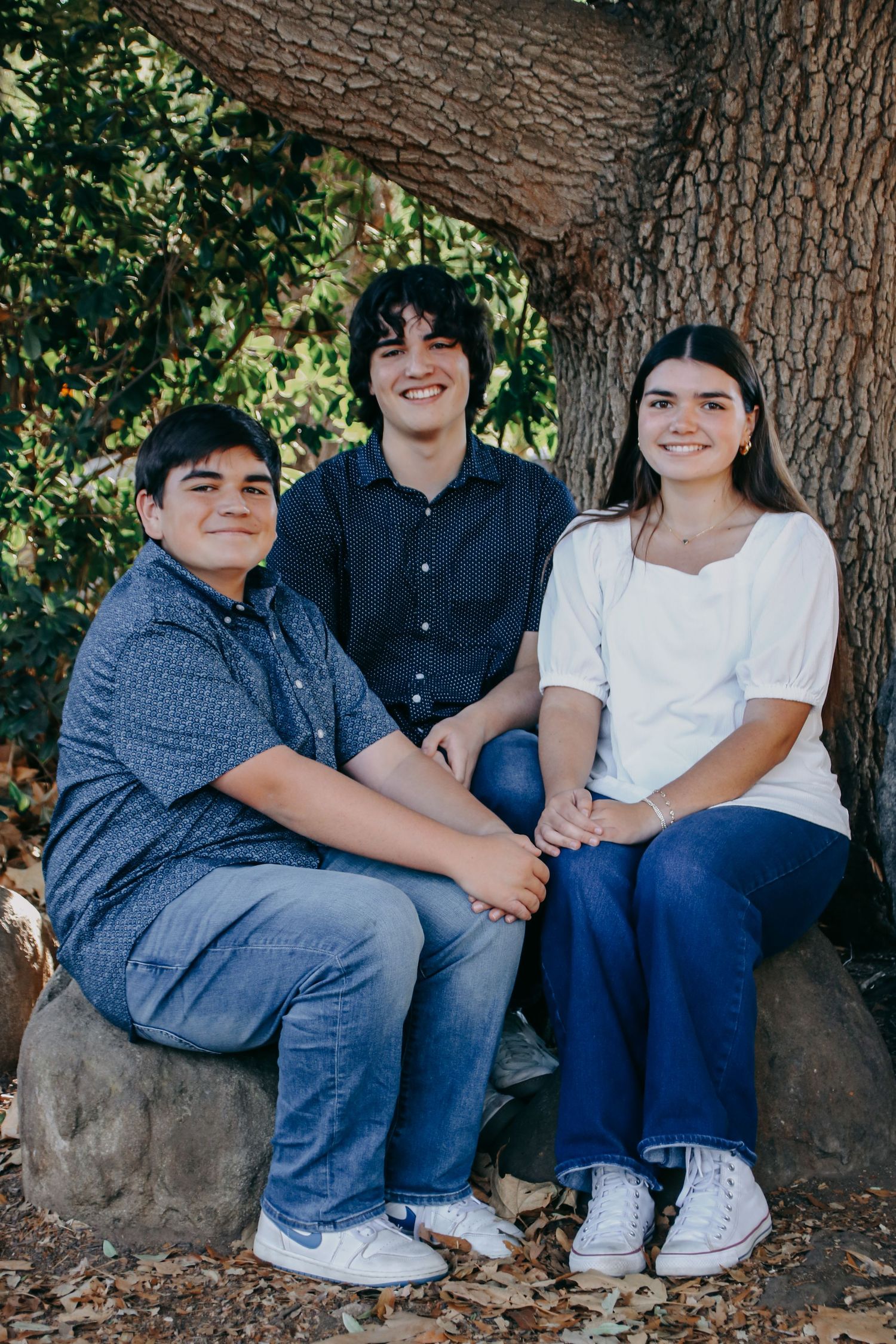 Three people sitting on a stone bench beneath a large tree in a casual outdoor photo session.