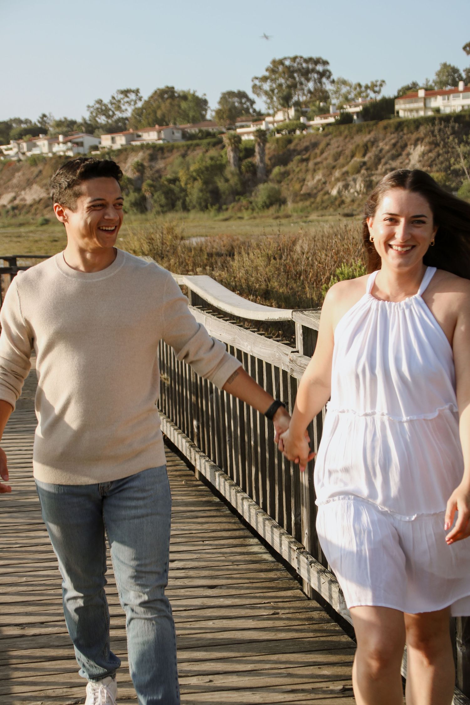 Two individuals share candid moments while walking along a wooden pathway by the shore.