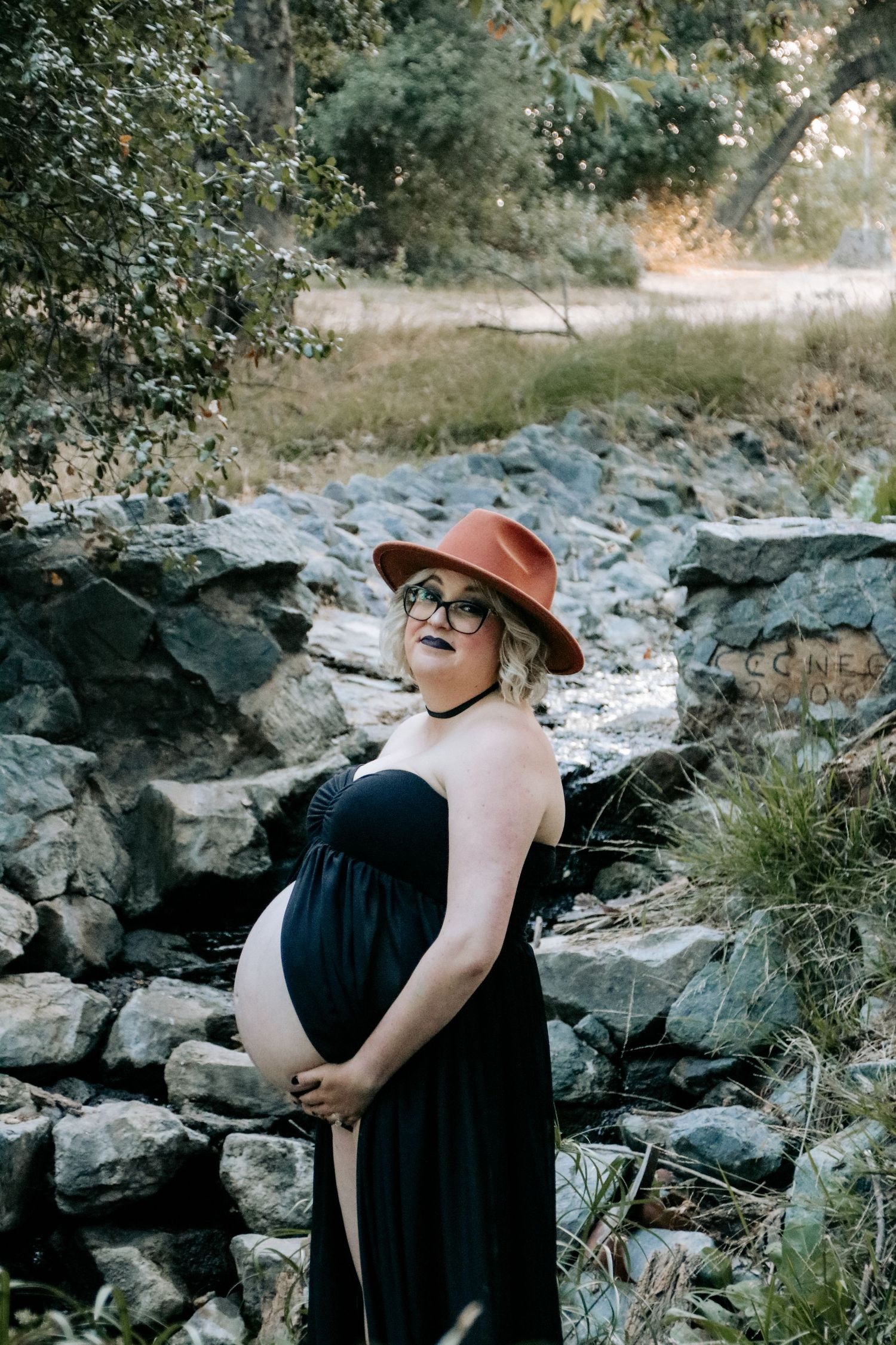 Outdoor maternity photos showing someone in a black dress and brown hat posing among rocky terrain and greenery.