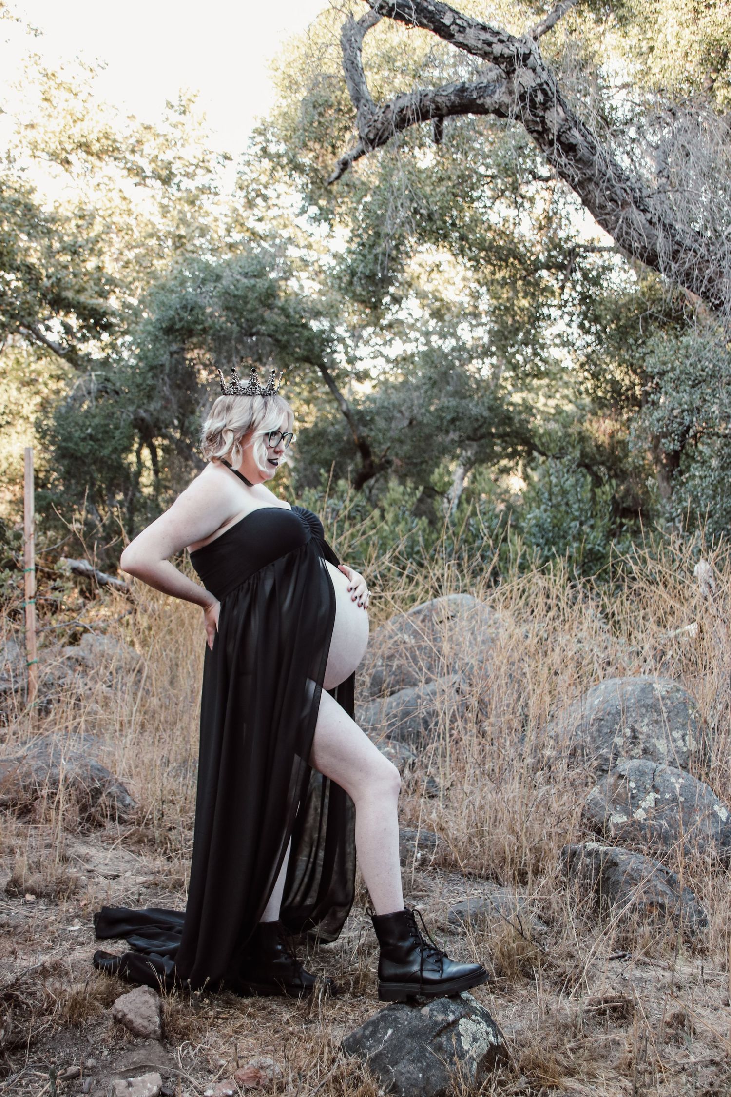 A maternity photo shoot in nature features a dramatic black flowing dress against a backdrop of dry grass and trees.