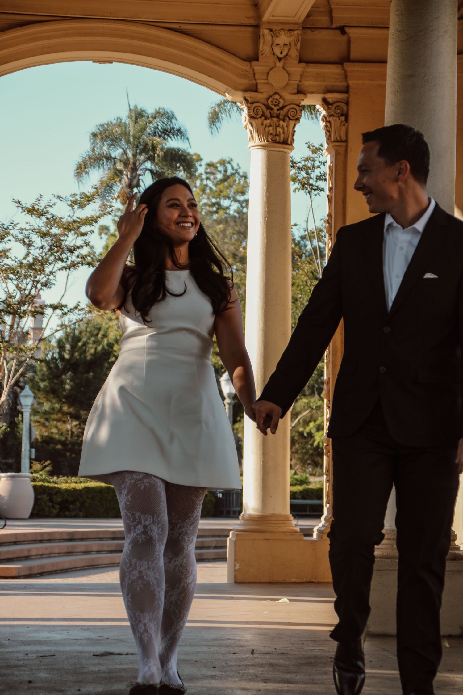 A happy couple walks hand in hand under decorative columns during golden hour.
