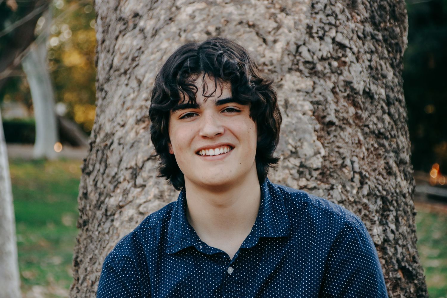 A person in a dark collared shirt poses casually while leaning against a tree on a sunny day.
