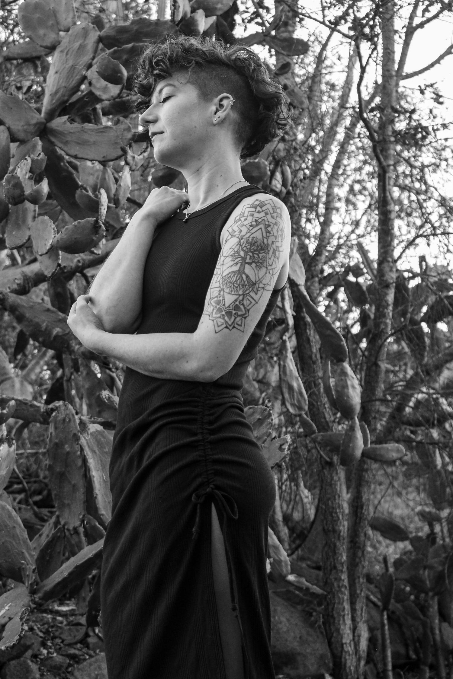 Figure in black dress poses artistically against tree in moody outdoor setting with cactus plants.