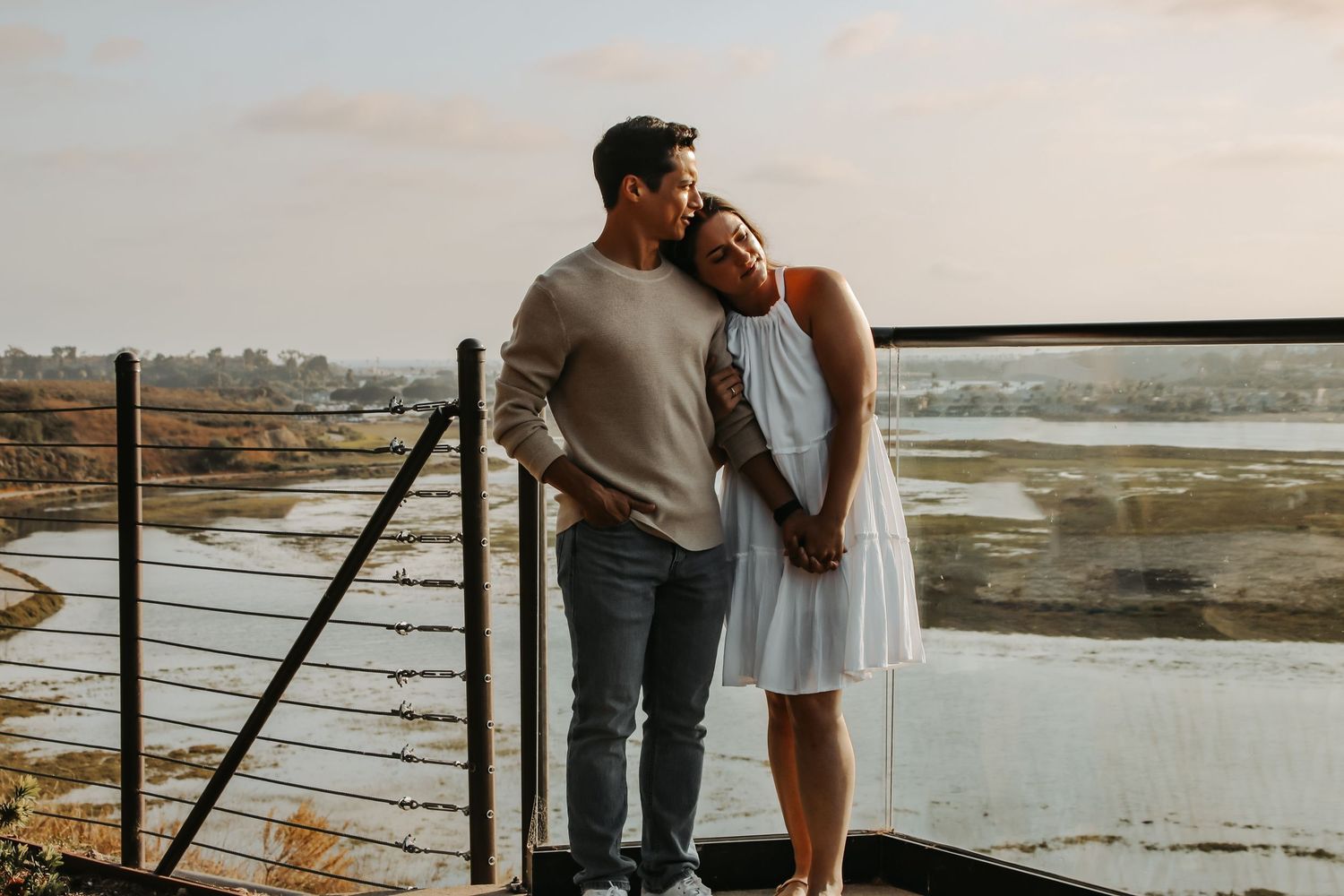 A couple embraces tenderly on a viewing platform overlooking a coastal marsh landscape.